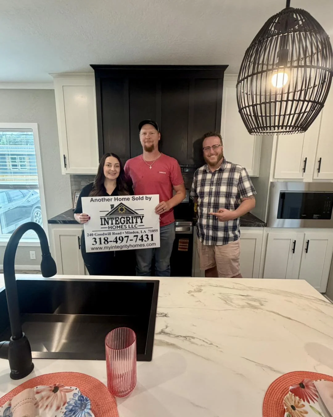 Three people standing in a kitchen, holding a sign that says 'Another Home Sold by Integrity Homes LLC, 240 Goodwill Road, Minden, LA 71055, 318-497-7431, www.myintegrityhomes.com.' The kitchen has black and white cabinets, a black range, and hanging