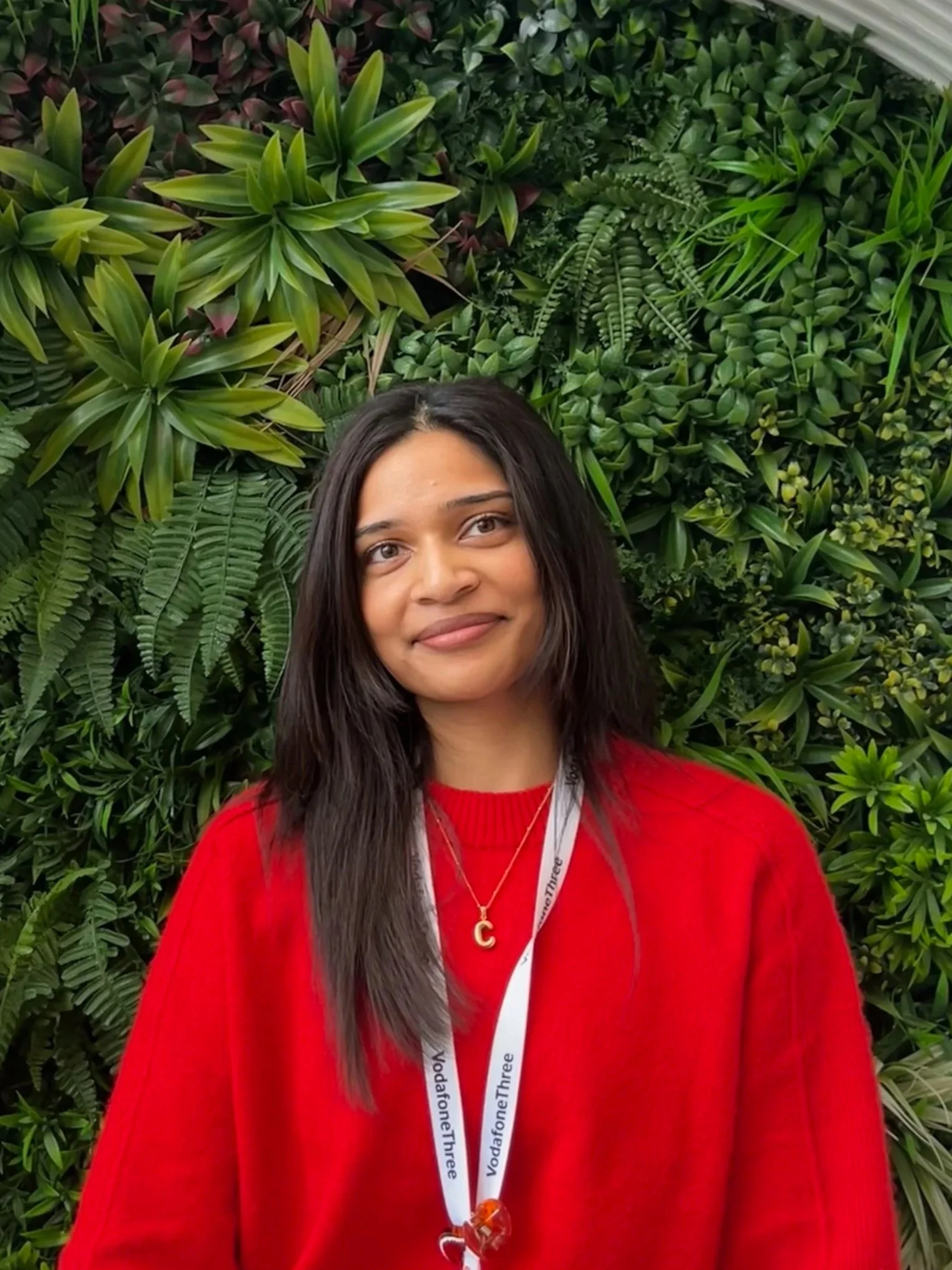 A woman with long dark hair wearing a red sweater and a necklace with a letter 'C', standing in front of a lush green leafy background, smiling at the camera.