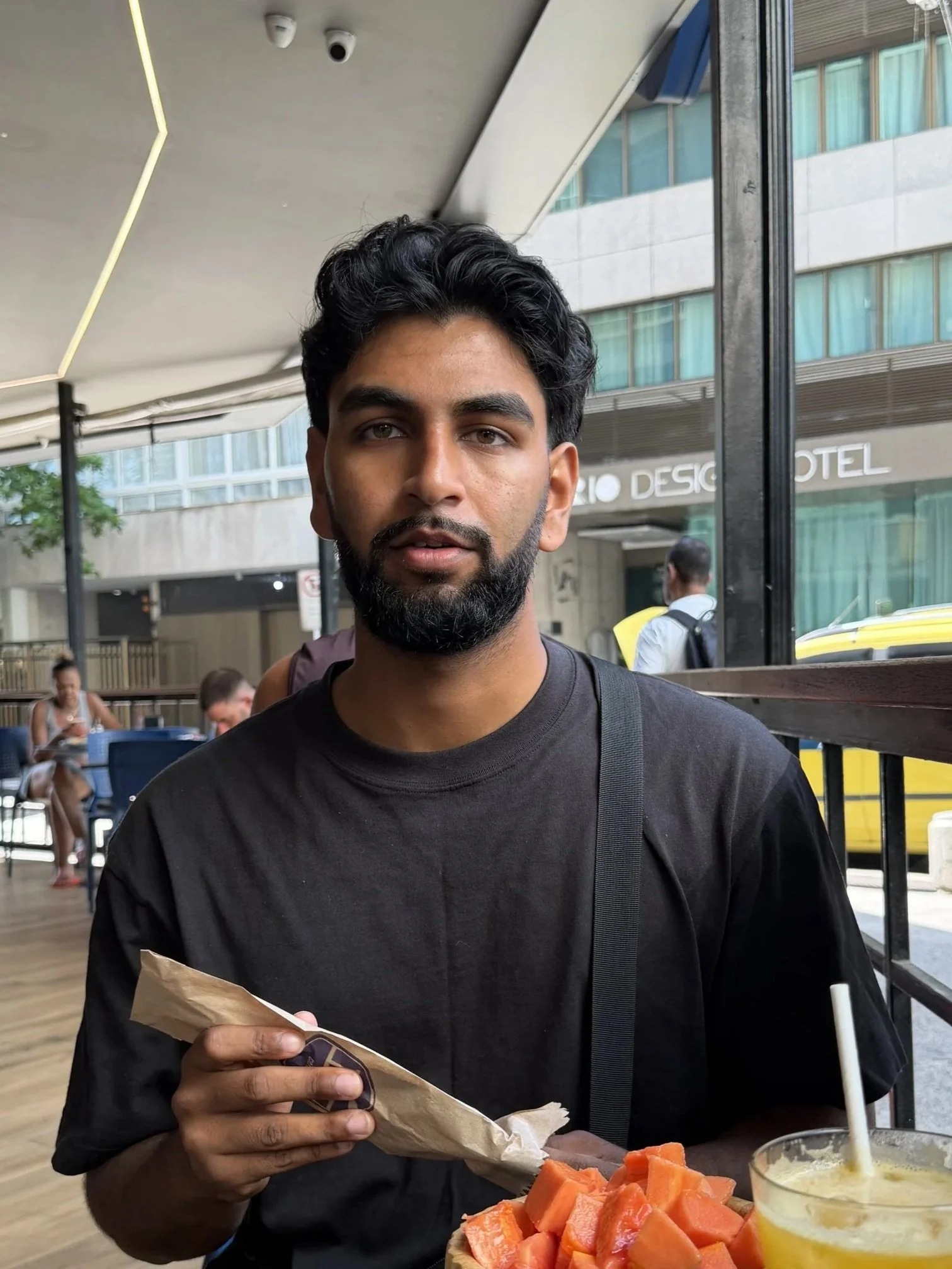 A man with dark hair and a beard wearing a black shirt, holding a partially wrapped food item, sitting at a table with chopped cantaloupe and a yellow drink with a straw in a busy outdoor dining area with modern buildings and people in the background.