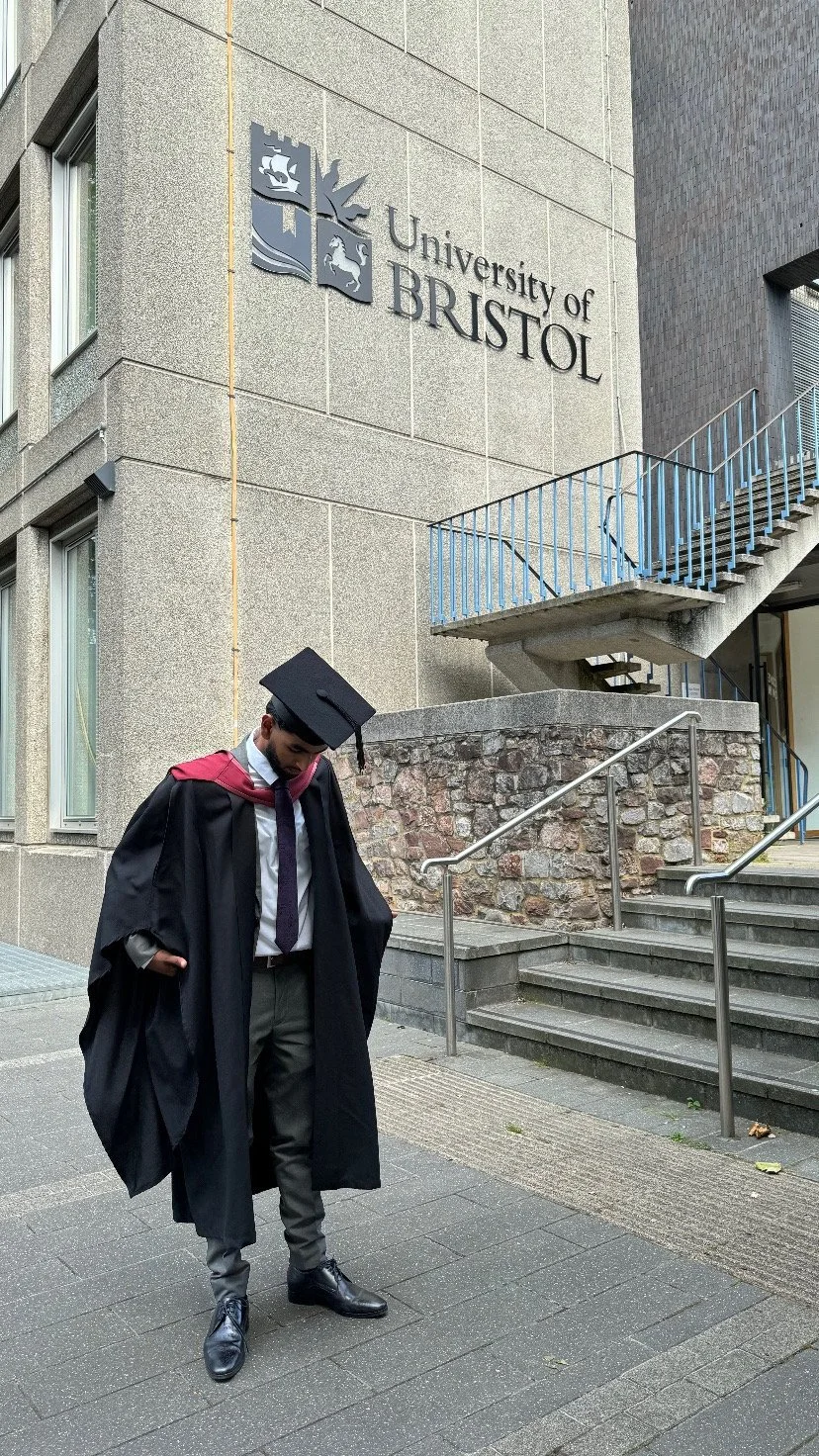 A graduate in cap and gown standing outside the University of Bristol building.