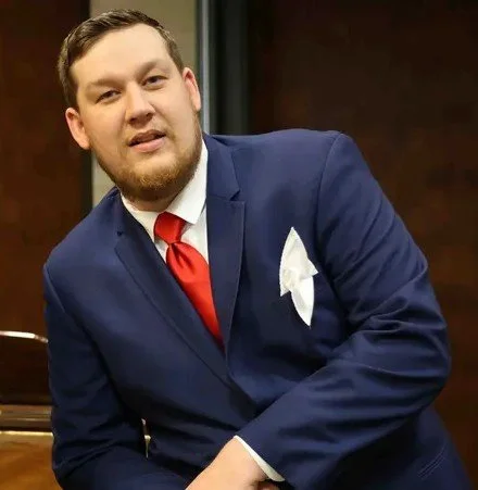 Man with short brown hair and a beard, wearing a blue suit, white shirt, red tie, and a white pocket square, sitting in a formal setting.