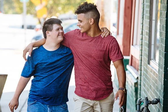 Two young men walking together on a sidewalk, smiling and looking at each other, with one arm around the other's shoulder.
