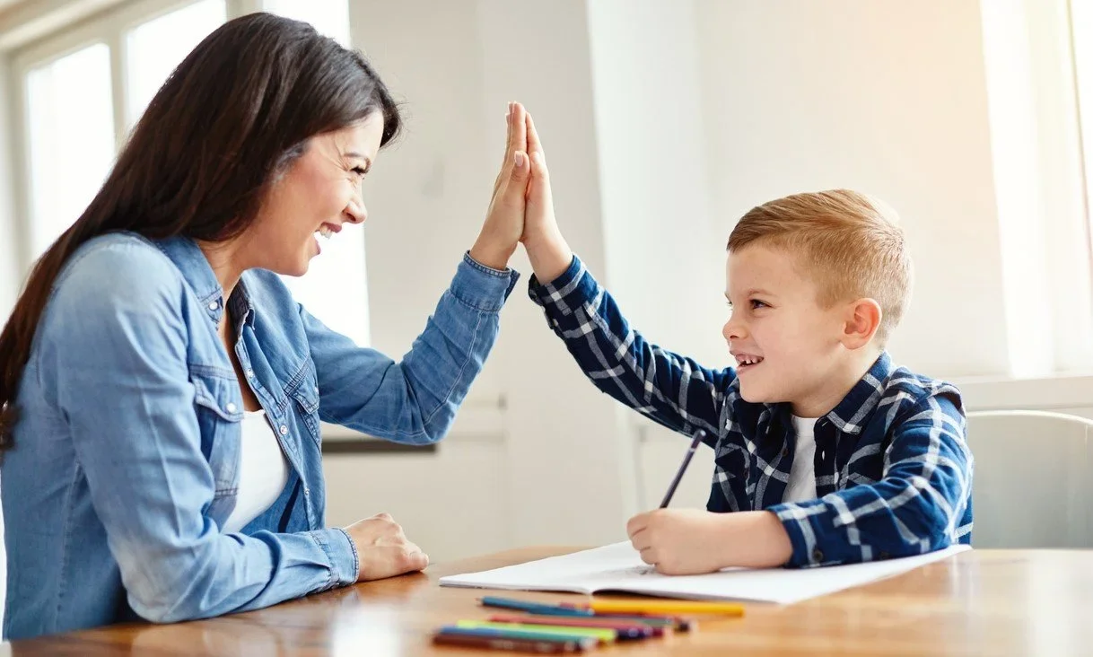 A woman and a young boy are giving each other a high five, sitting at a desk with a notebook and colored pencils. They are smiling and appear happy.