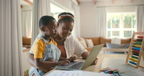 A woman and a young girl sitting at a table, looking at a laptop together in a bright living room.