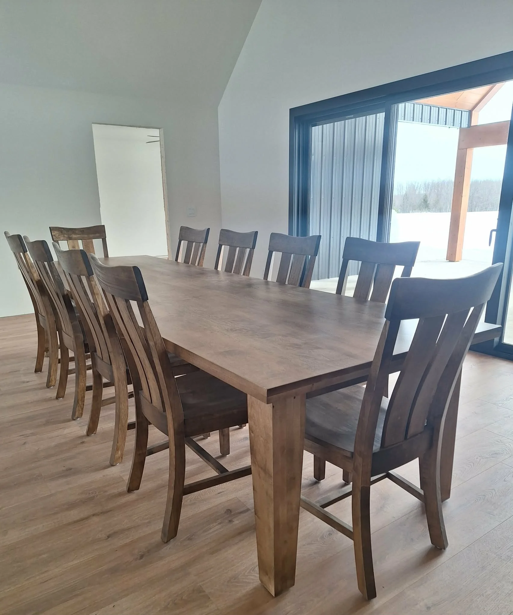 A wooden dining table with ten matching chairs inside a room with wooden flooring, large windows, and white walls. Outside the window, there is a view of snow and trees.