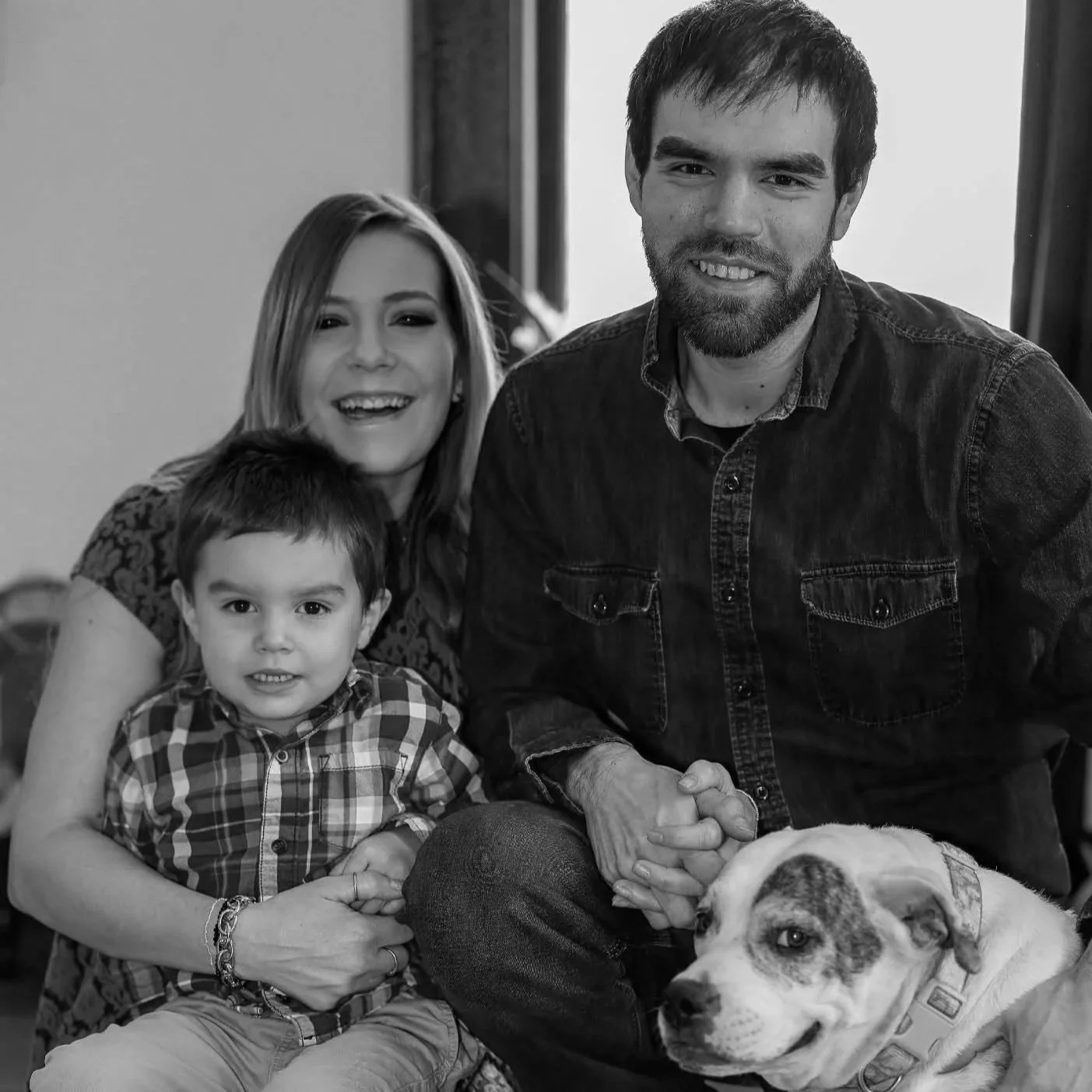 A black and white photo of a family of three with their dog, sitting indoors. The woman has shoulder-length hair, and the man has a beard. The boy in front is wearing a plaid shirt. The dog is lying down in front of them, wearing a bandana.