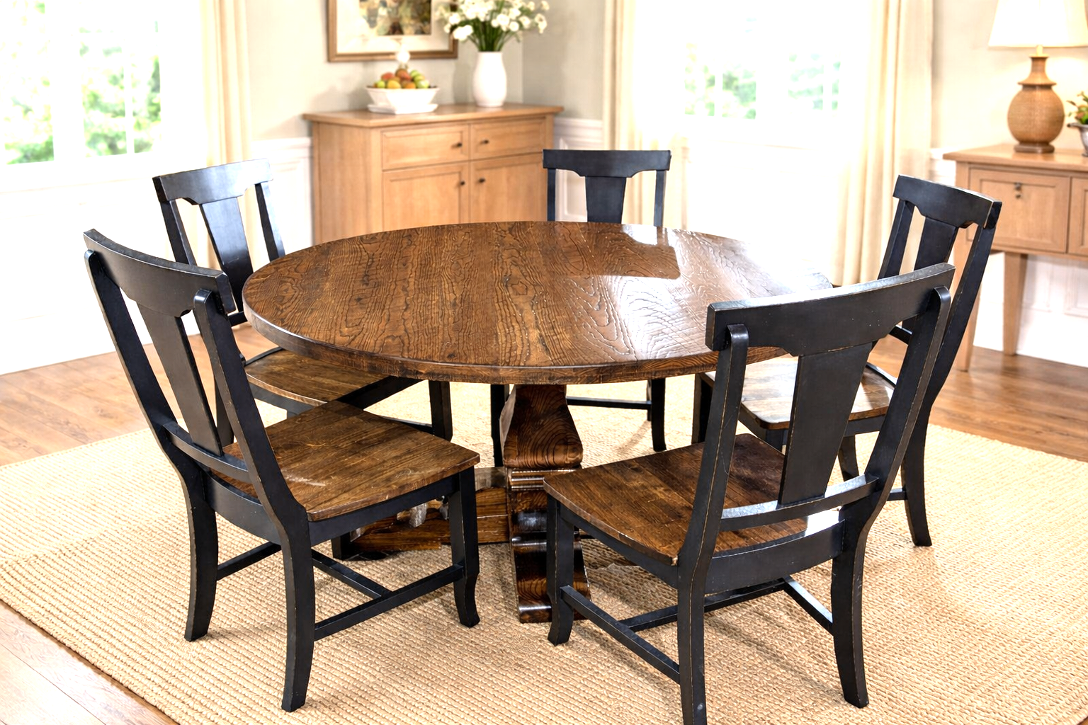 A wooden dining table with five black chairs arranged around it in a bright, sunlit room with large windows, light-colored curtains, and wooden furniture.
