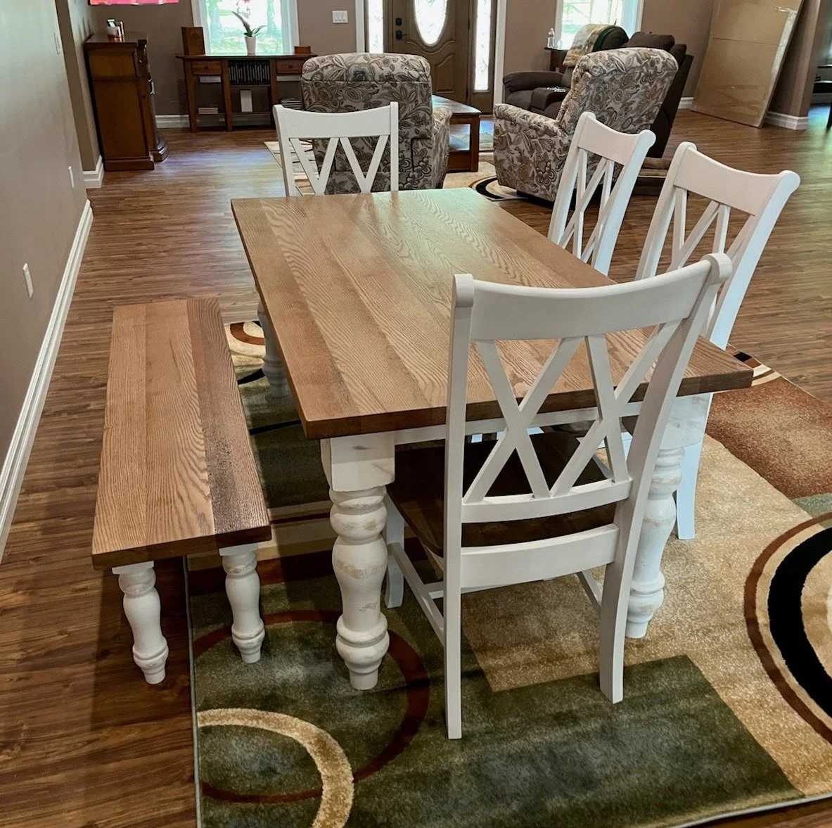 A wooden dining table with white legs and six chairs, four with white frames and two with wooden frames, in a living room area with patterned armchairs, a wooden sideboard, and a brown armchair in the background.
