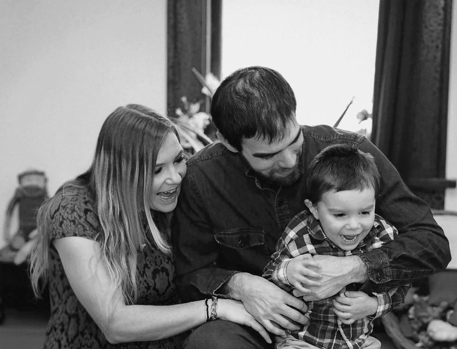 A black-and-white photo of a family playing together indoors. A young woman, a man, and a young boy are happily engaged, with the woman and man smiling and the boy laughing. The woman has long hair and is wearing a patterned top. The man has short hair and a beard, wearing a casual shirt. The boy is wearing a checkered shirt, and they are all holding or near each other, sharing a joyful moment.