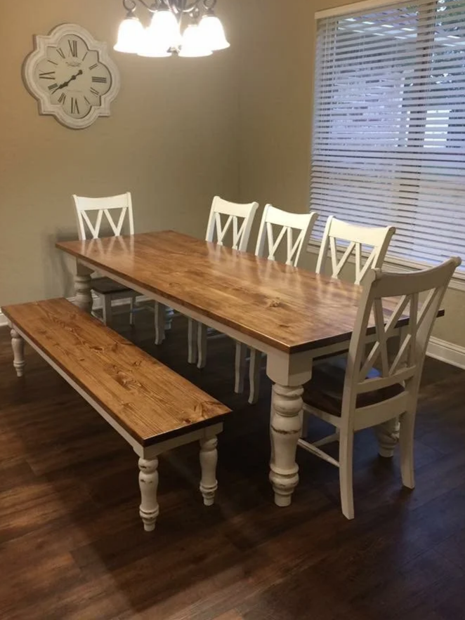 A wooden dining table with six white chairs and a wooden bench in a dining room with dark hardwood floors, a wall clock, and a window with blinds.