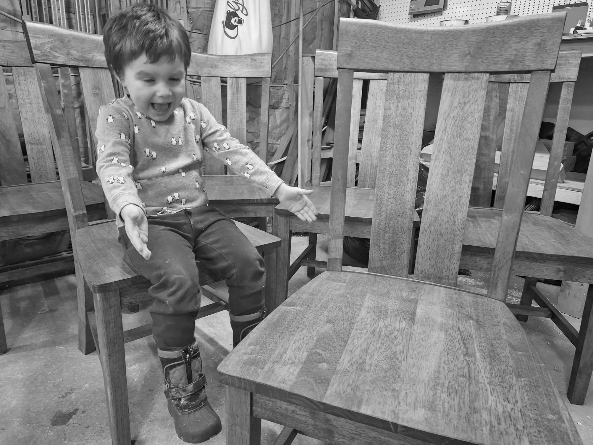 A young child sitting on a wooden chair, wearing a patterned sweater and boots, with empty hands raised, in a workshop or wood shop environment.