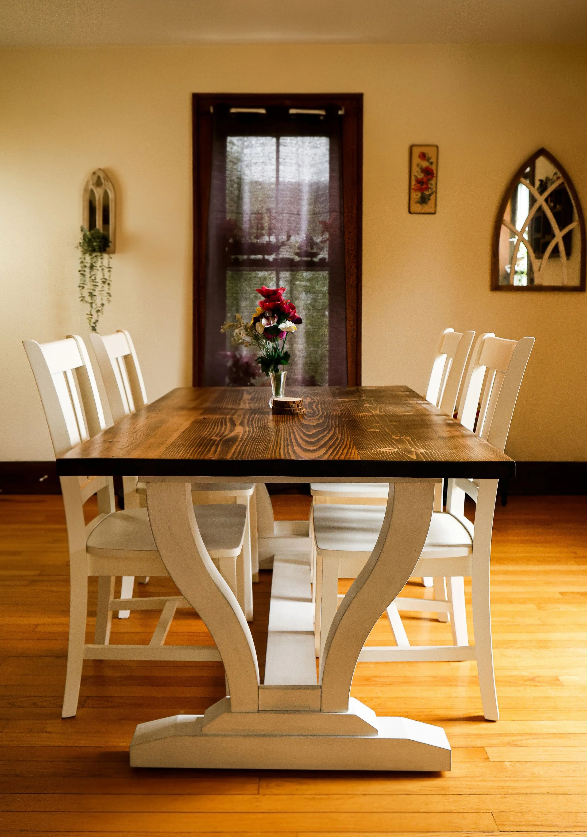 A dining room with a wooden table and six white chairs, a glass vase with red, white, and pink flowers on the table, a window with brown trim and sheer curtains at the back, and walls decorated with framed artwork and a mirror.