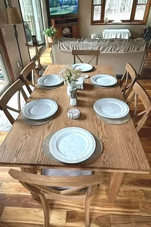 Wooden dining table set with six white plates, salt and pepper shakers, a small basket, and a floral centerpiece in a cozy home dining area.