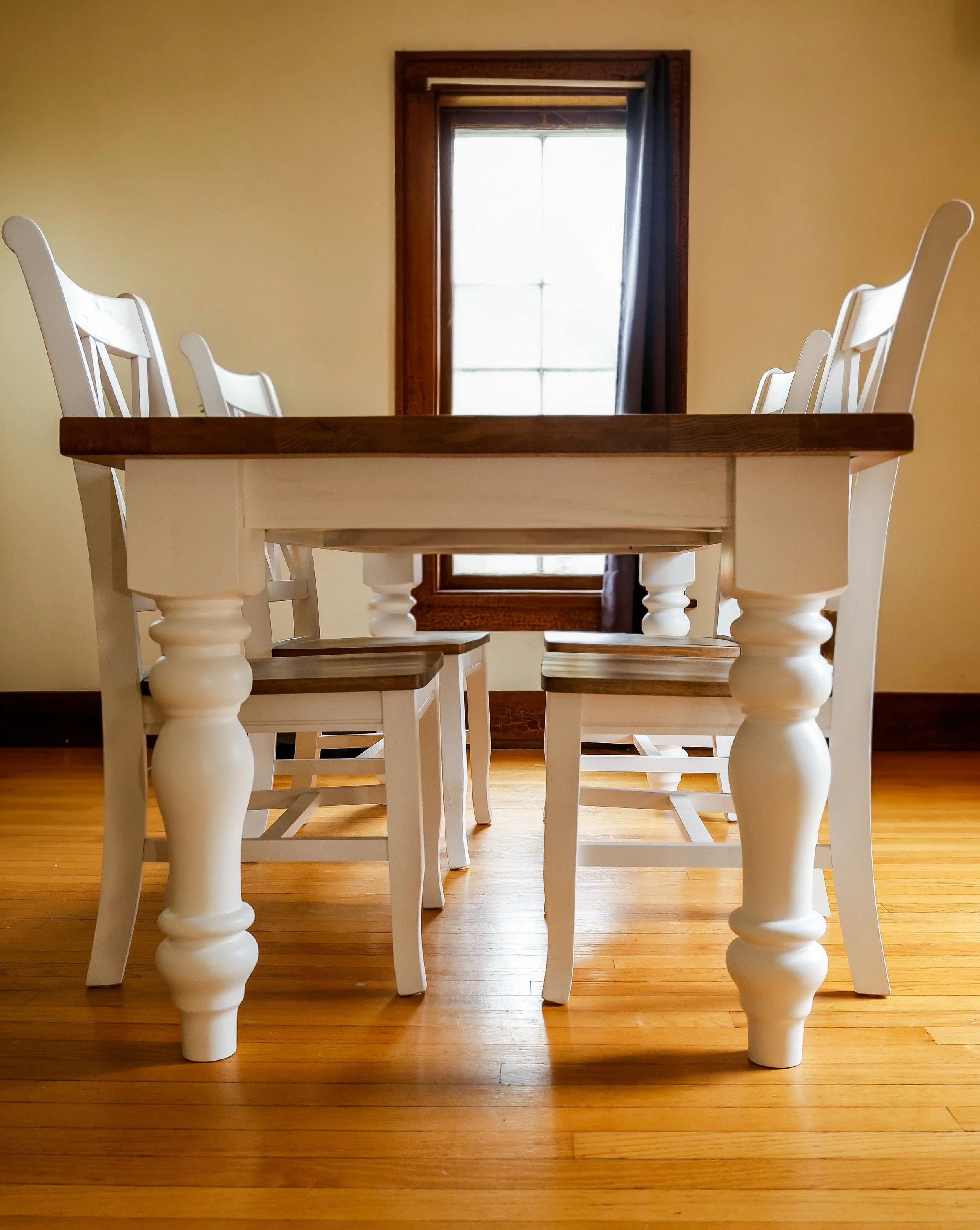 White wooden dining table with six matching chairs on a oak floor, a window with dark curtains in the background