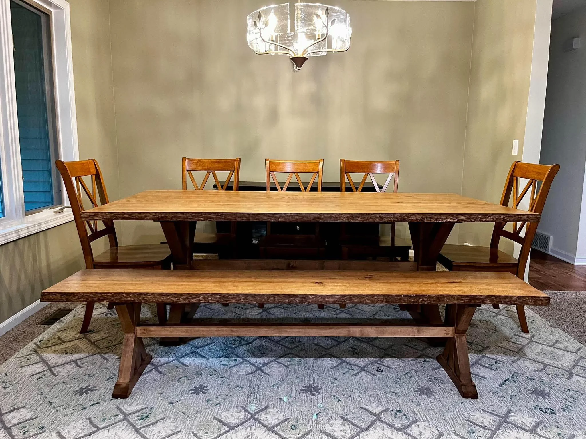A wooden dining table with six matching chairs in a dining room, featuring a large chandelier hanging from the ceiling and a window on the left side of the room.