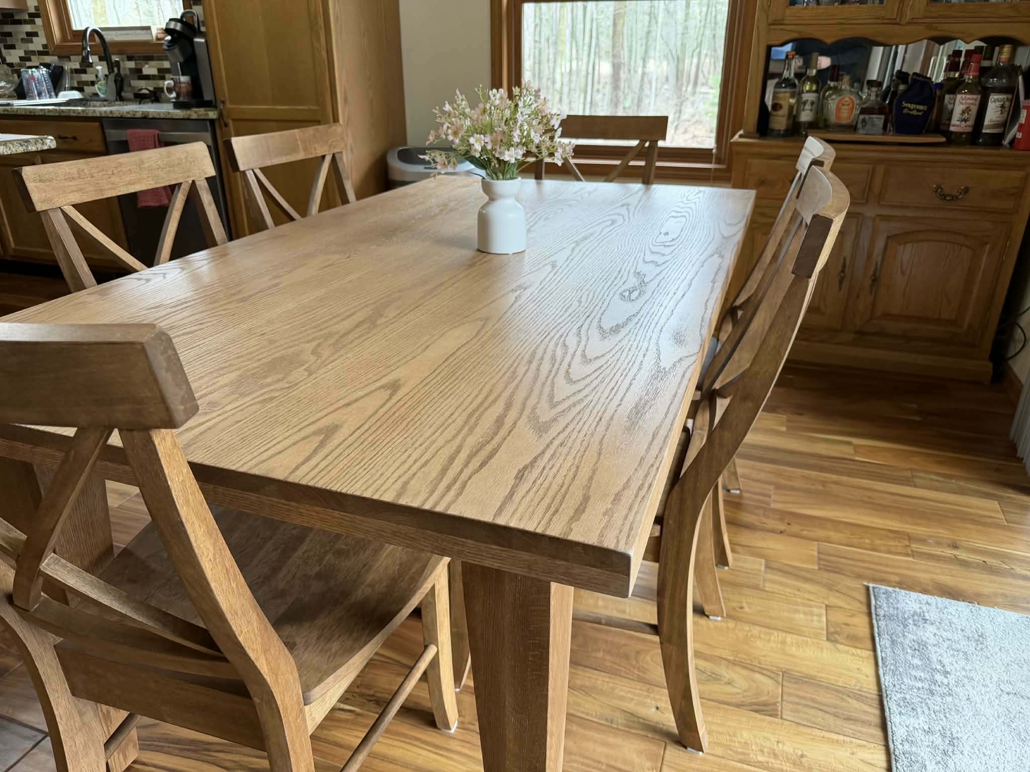 A dining room with a large wooden table and six matching chairs. A white vase with pink and white flowers is on the table. In the background, there is a wooden cabinet with bottles of alcohol and a window showing trees outside.