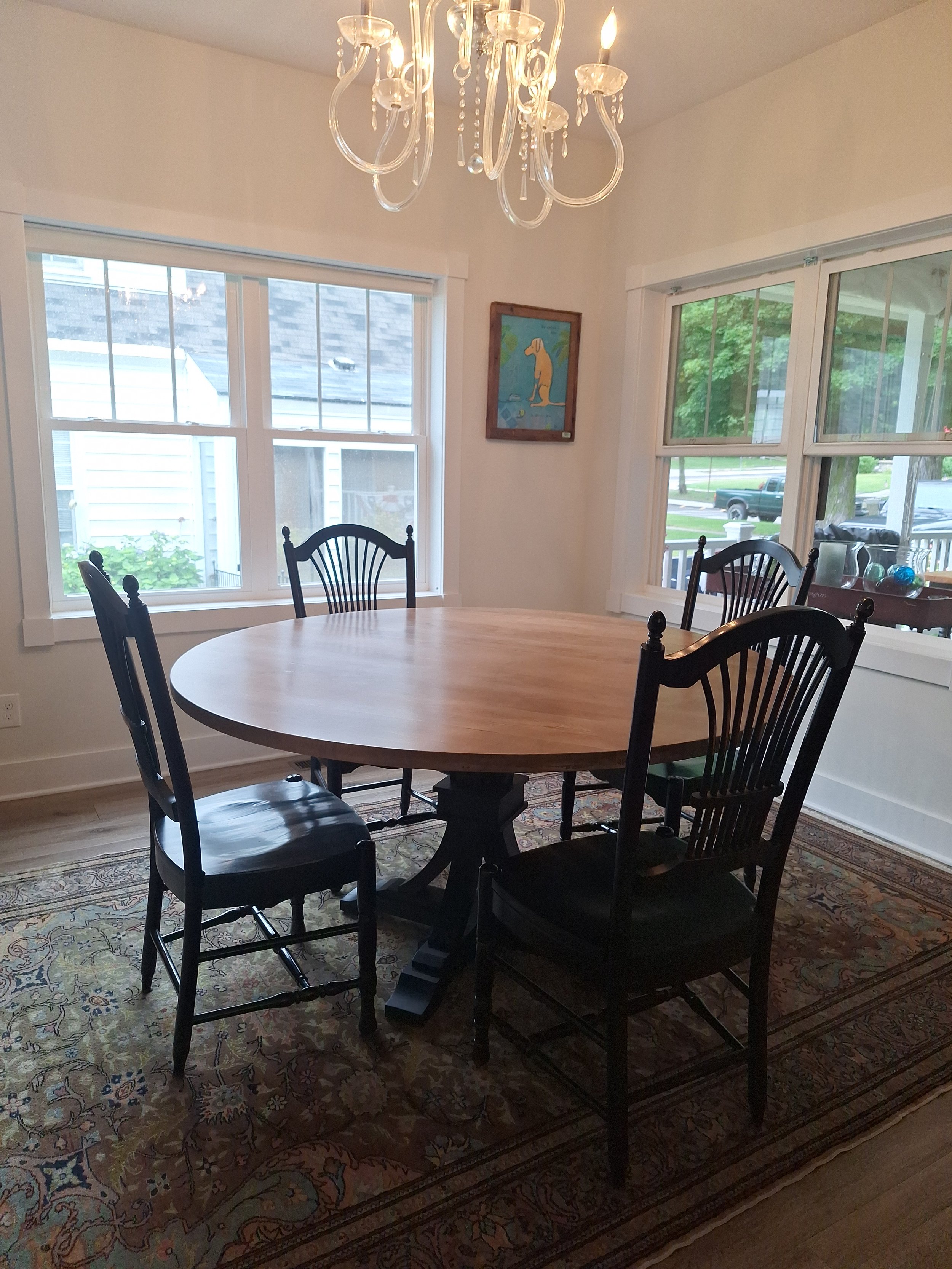 A dining room with a round wooden table, six black chairs, a patterned area rug, large windows, a framed dog-themed artwork, and a chandelier overhead.