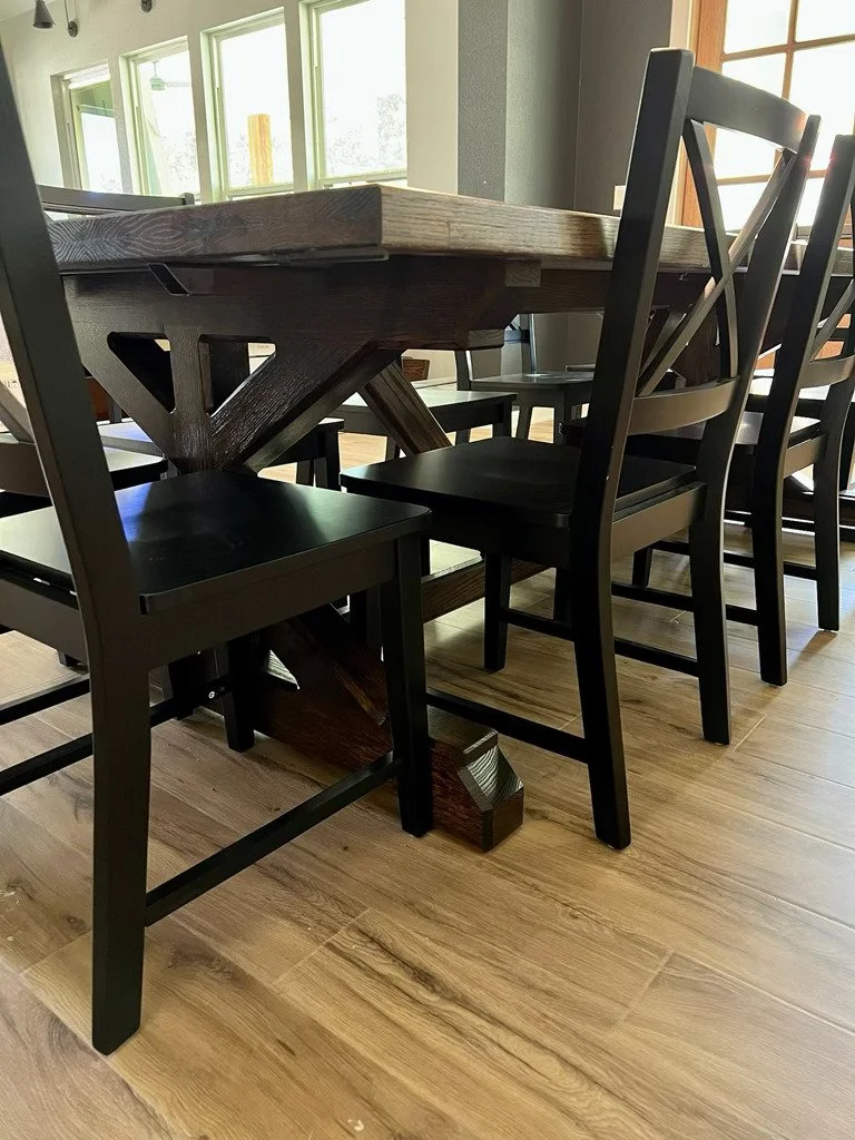 Close-up of a wooden dining table with black chairs around it in a well-lit room with windows.