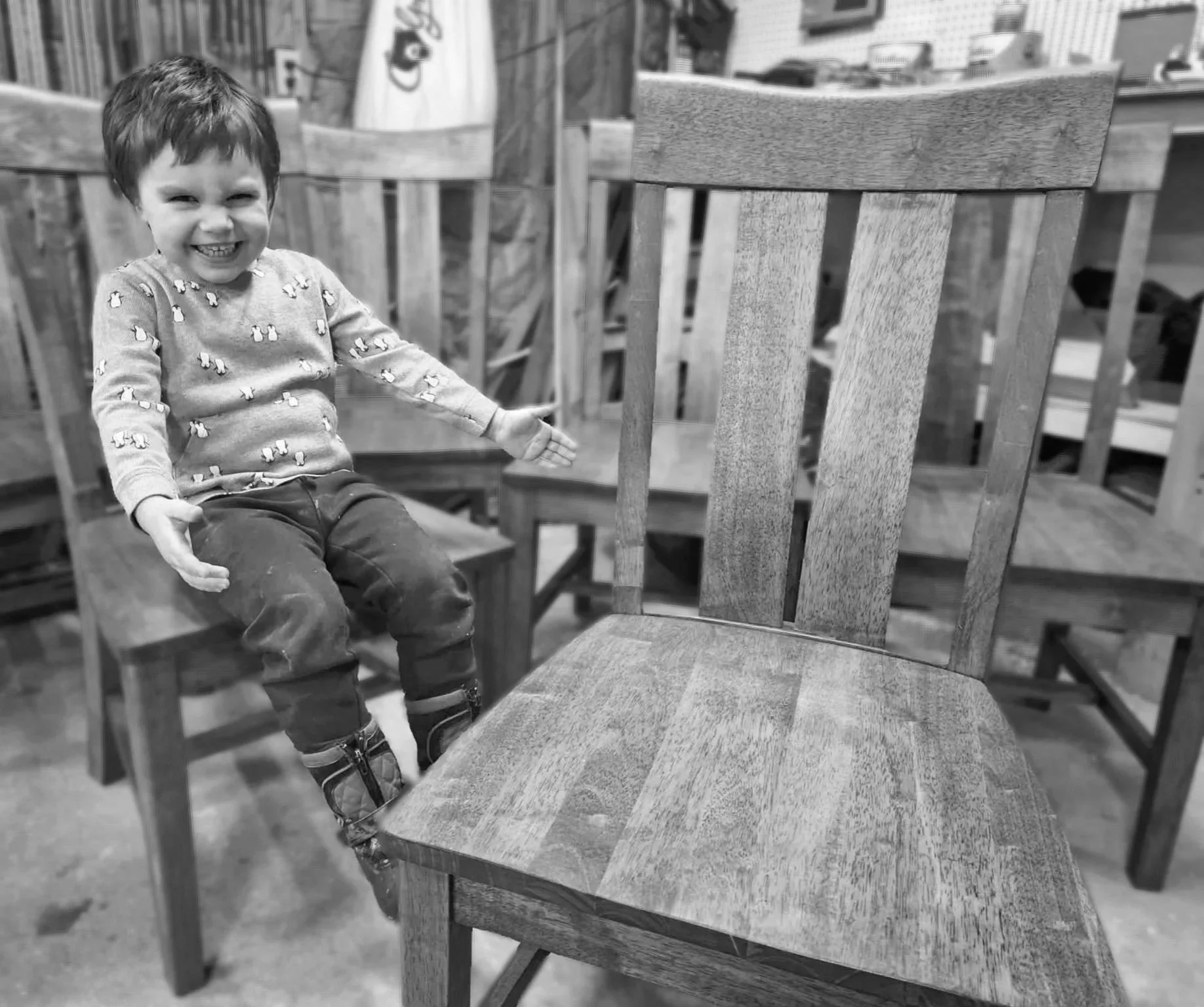 A young boy sitting on a wooden chair with a big smile, wearing a sweater with small animal patterns and dark pants, in a store or workshop with wooden furniture and various objects in the background.