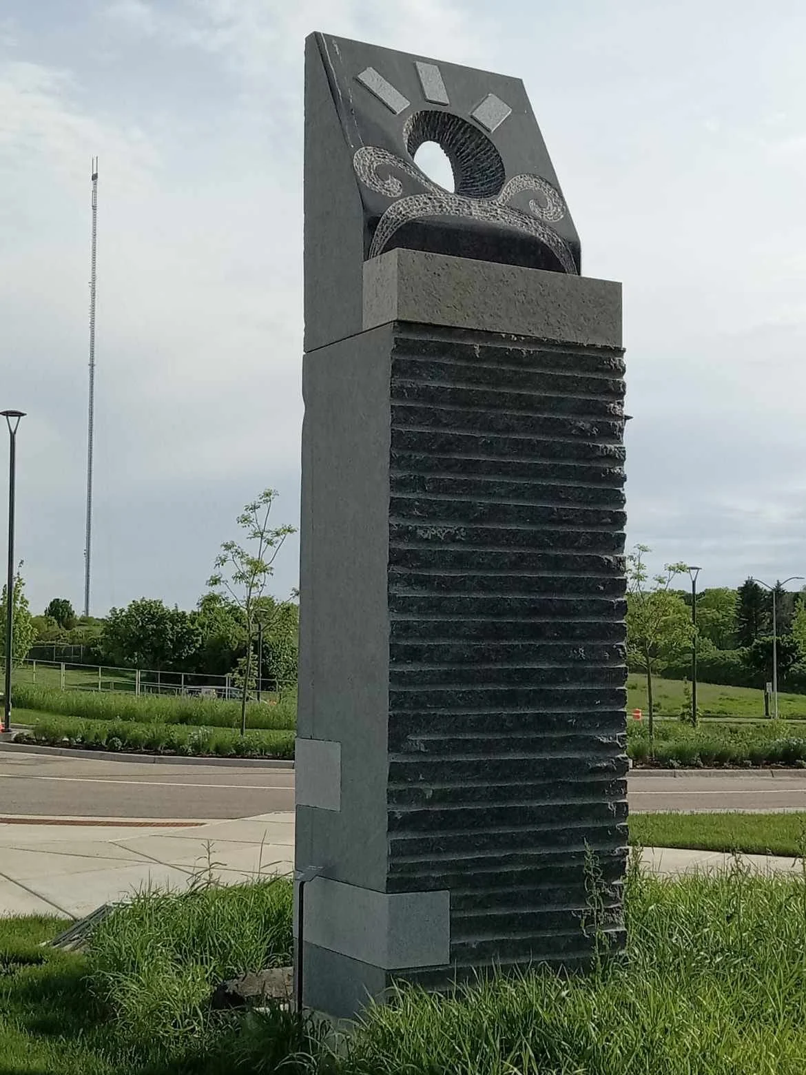 Outdoor monument with a modern abstract design, featuring a black and gray color scheme, with a circular opening at the top and decorative swirling patterns, mounted on a ribbed rectangular base, set in a park-like area with trees and grass.