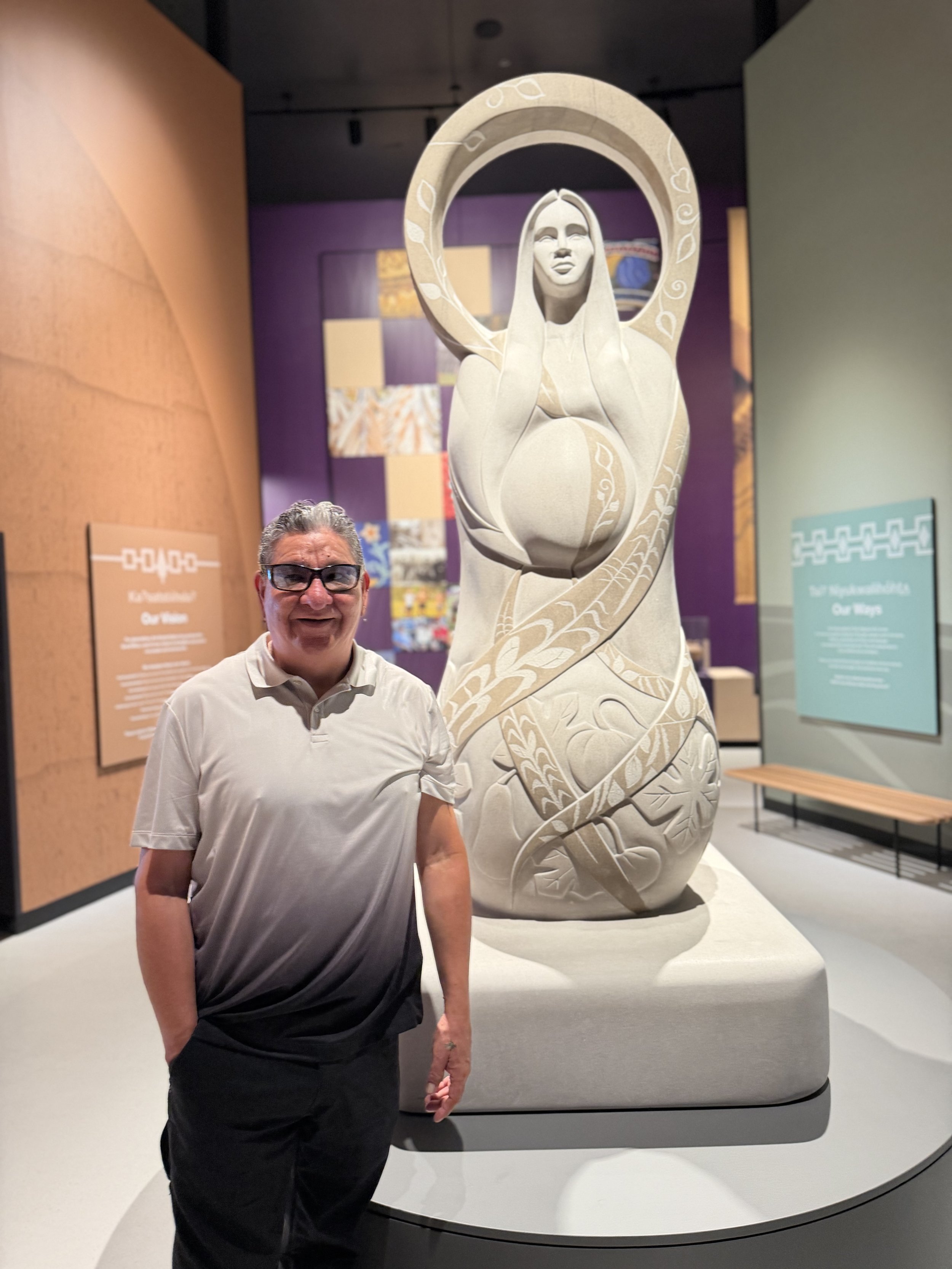 A man with gray hair, glasses, and a white polo shirt standing in front of a sculpture of a woman with long hair, who is holding a circular object around her head. The background features colorful artwork and informational signs.