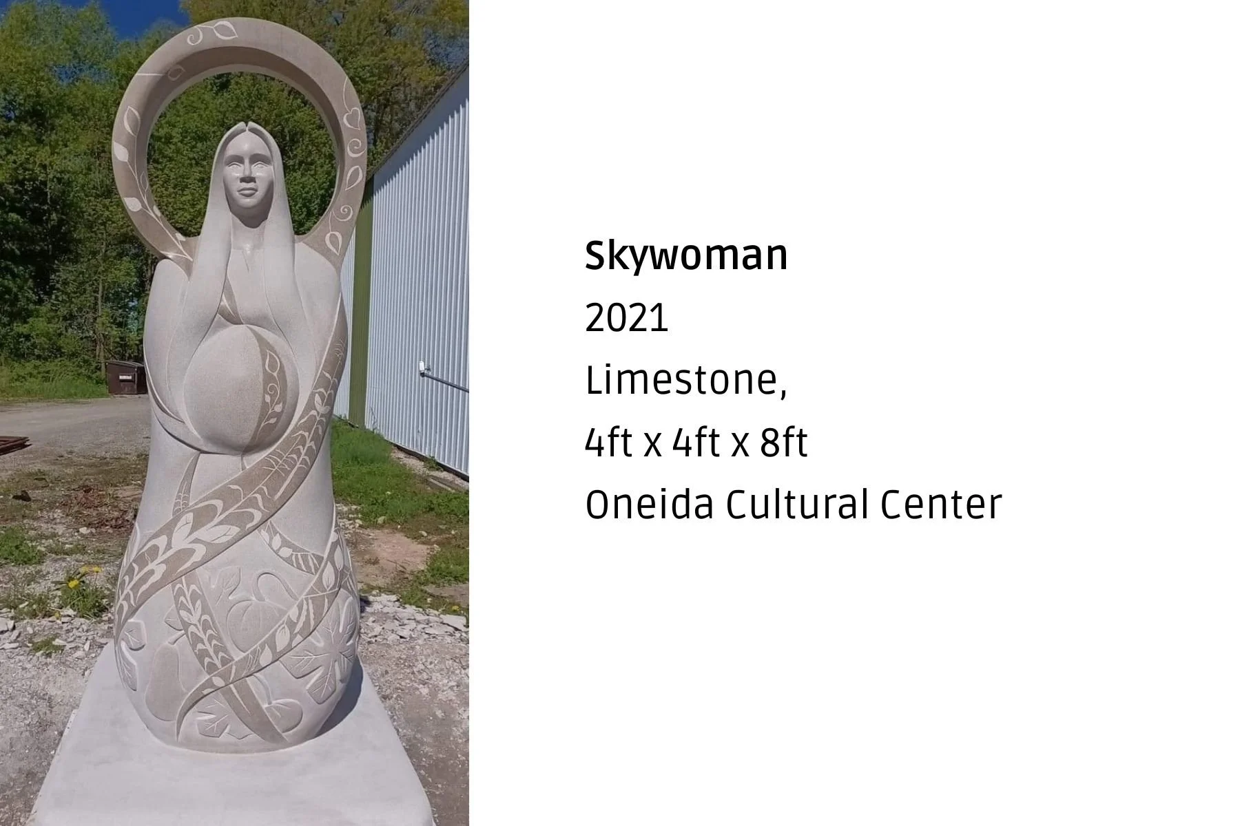 A white limestone sculpture of a woman with long hair and a halo featuring leaf and heart patterns, standing outdoors with trees and a metal building in the background.