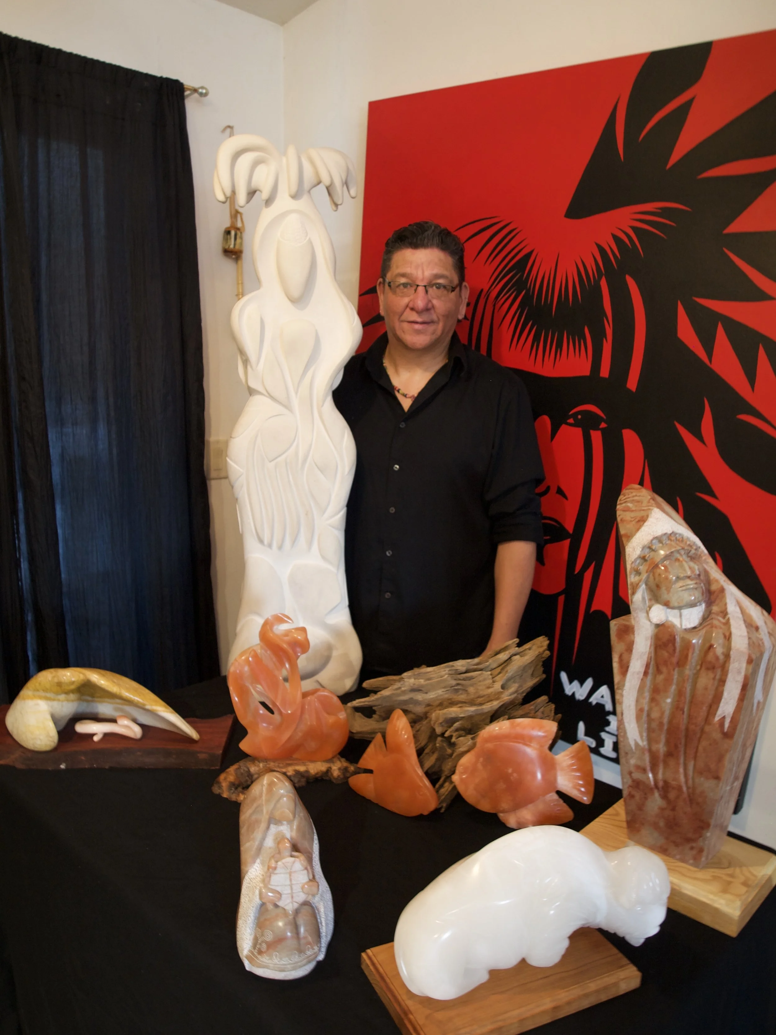 Display of carved and polished stone sculptures on a black table, including animal shapes and abstract forms, with a man standing behind them against a red and black artwork backdrop.