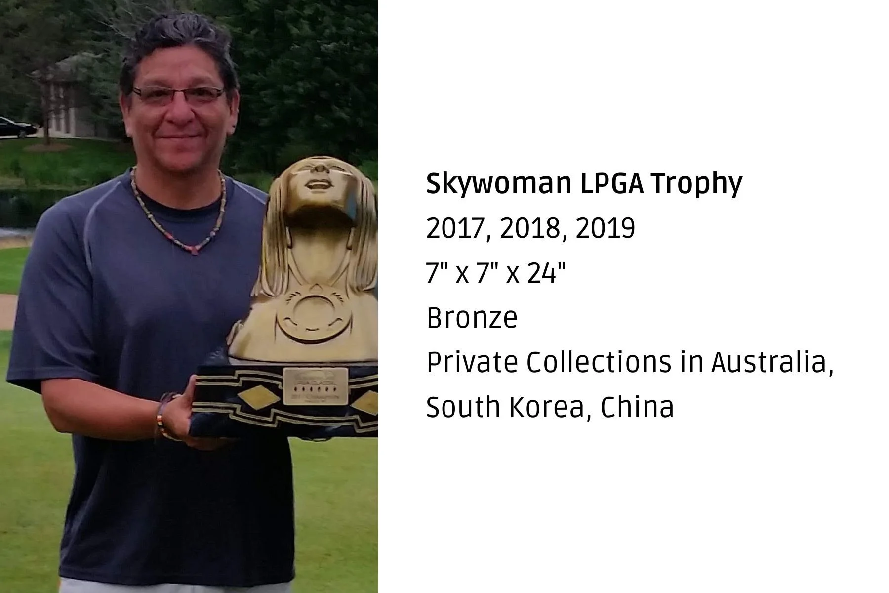 A man wearing glasses and a dark blue shirt holding a trophy. The trophy is shaped like a woman with long hair and a big smile, holding a golf club. There are trees and a water body in the background.
