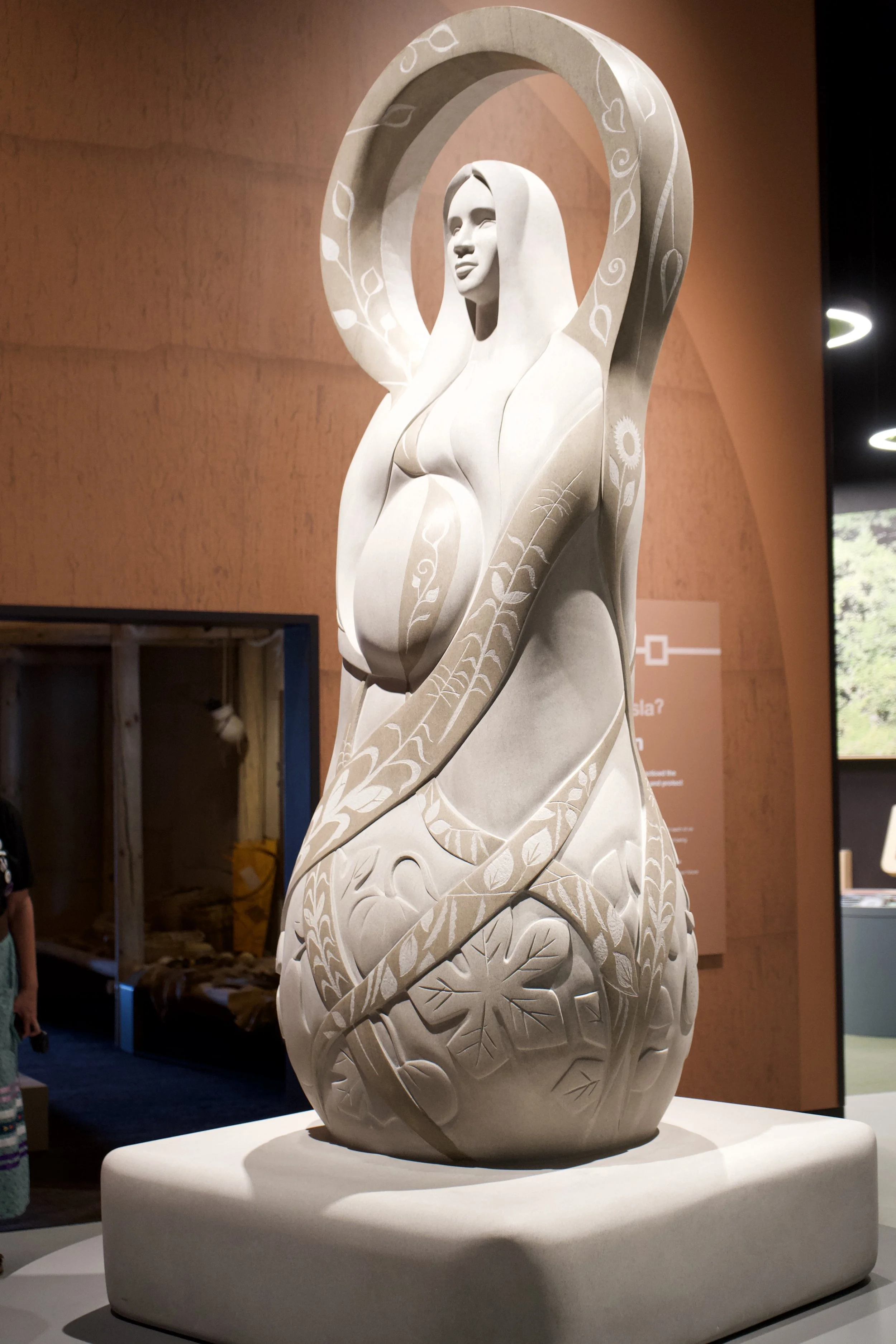 A white sculpture of a woman with long flowing hair, surrounded by intricate floral and leaf patterns carved into the surface, displayed on a white pedestal.