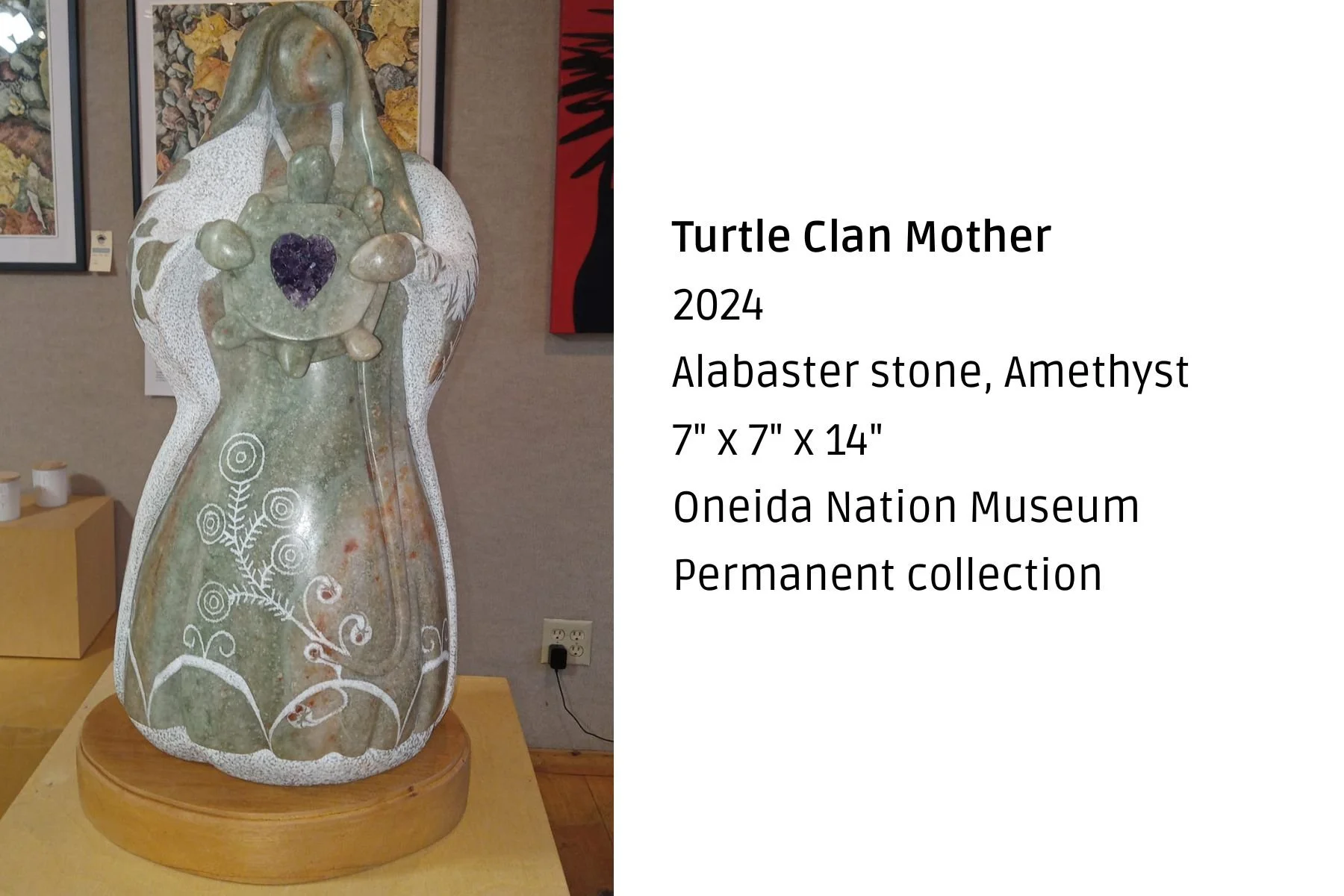 A carved alabaster stone sculpture titled 'Turtle Clan Mother' displayed on a wooden stand. The sculpture features intricate white patterns and a prominent amethyst stone in the center of the turtle's shell, with a figure of a mother holding a child 
