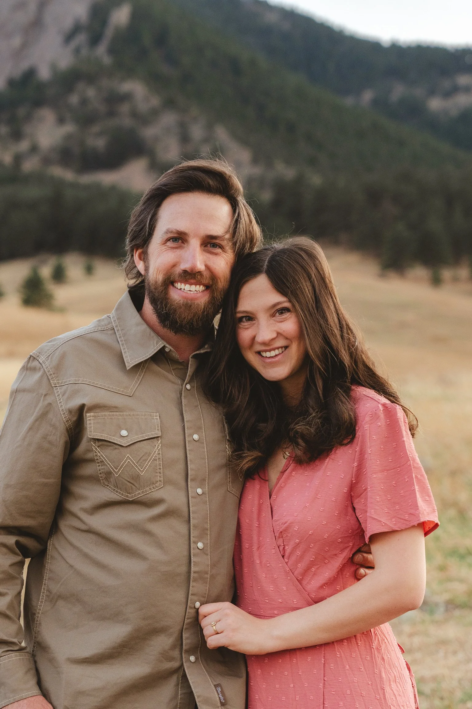 A smiling man and woman standing outdoors in a field with mountains in the background, hugging each other.