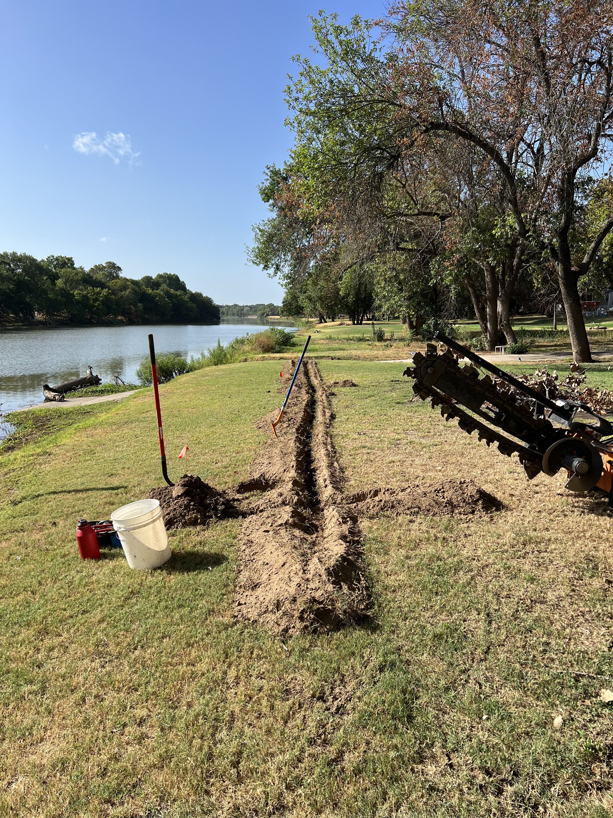 A riverside park with a clear blue sky, trees on the right side, and a partially dug trench in the grass with construction tools nearby.