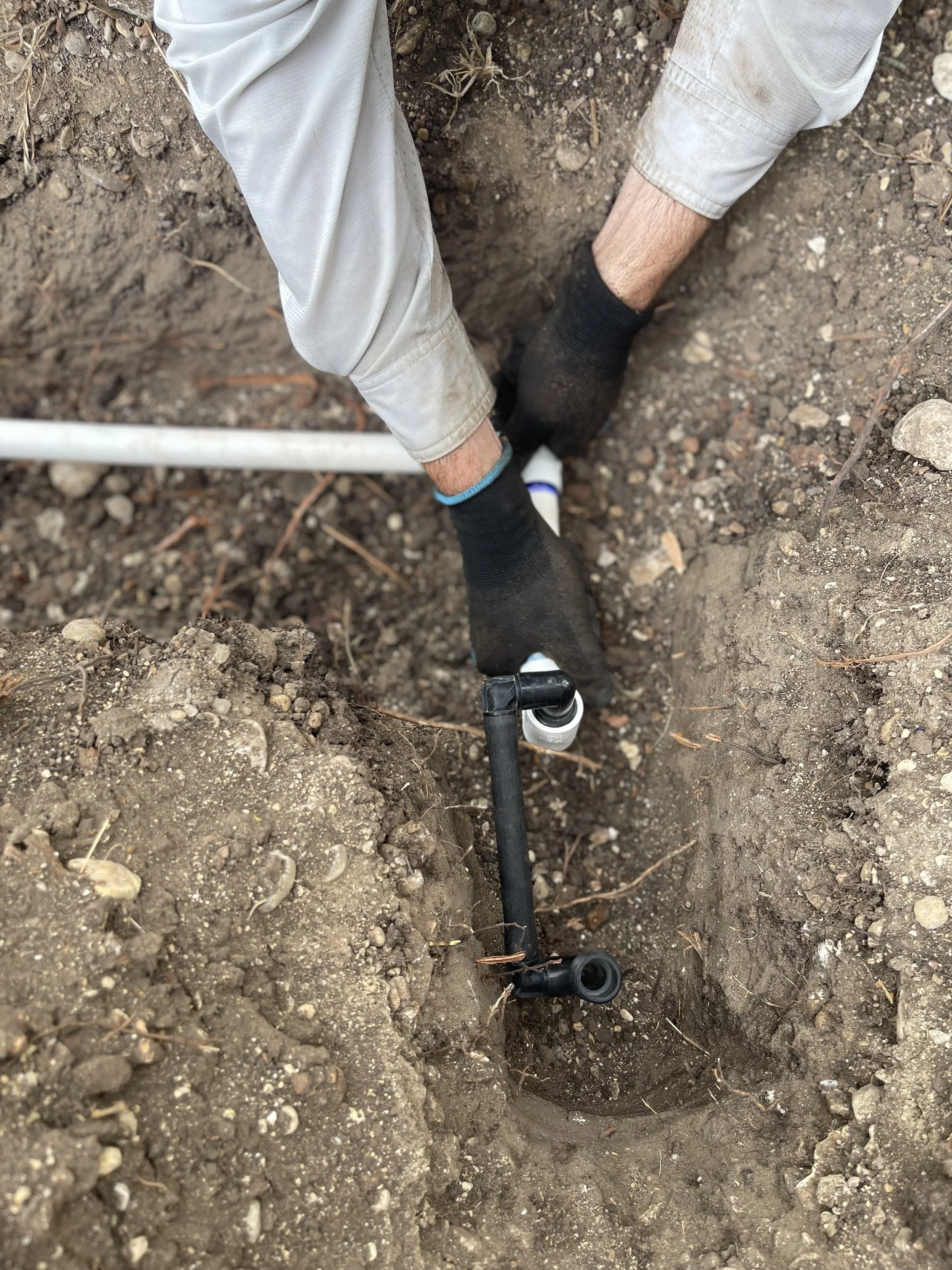 Close-up view of a person working with a black plastic pipe in a dirt trench, wearing black gloves and light-colored pants.