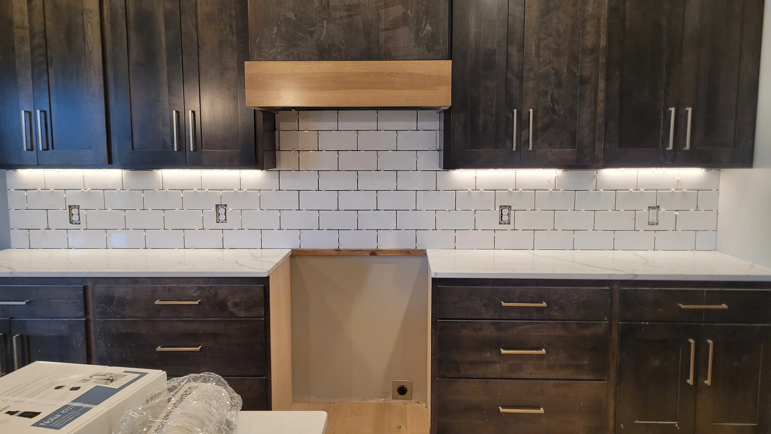 Kitchen with dark cabinets, white subway tile backsplash, and a marble countertop. Incomplete space with an empty cabinet area and multiple electrical outlets.