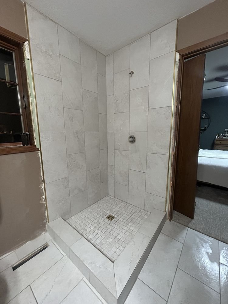Empty walk-in shower with beige marble tiles, small square mosaic tiles on the shower floor, and a central drain, adjacent to a bedroom with brown door frames and dark-painted walls.