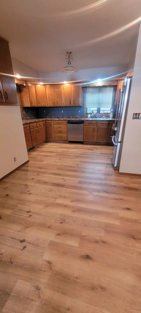 Empty kitchen with wooden cabinets, stainless steel dishwasher, refrigerator, and a window with blinds, hardwood floor, and a ceiling light fixture.