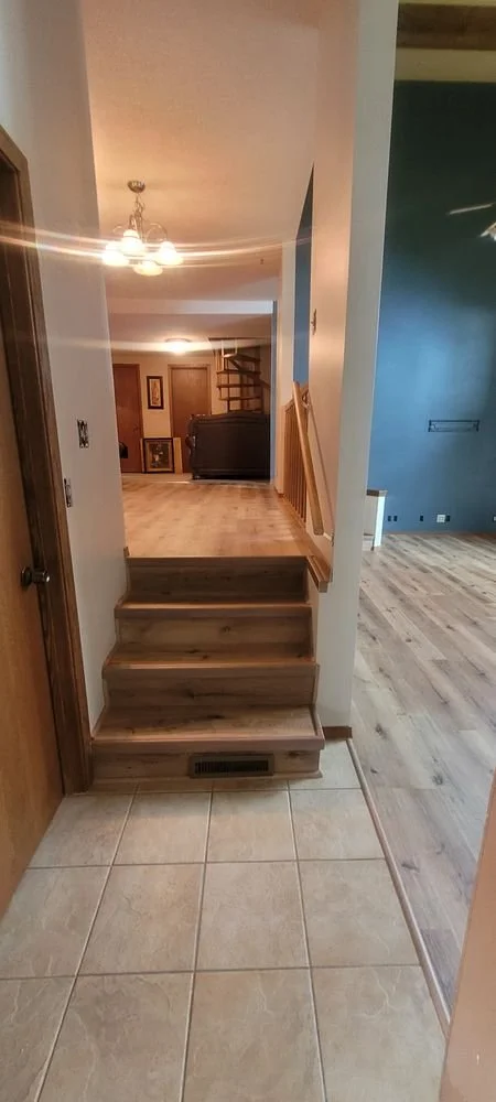 Interior view of a house featuring a small set of three wooden stairs leading up to a tile and wood floor area. The background shows a living space with a staircase, a dark-colored couch, framed pictures on the wall, and warm lighting.