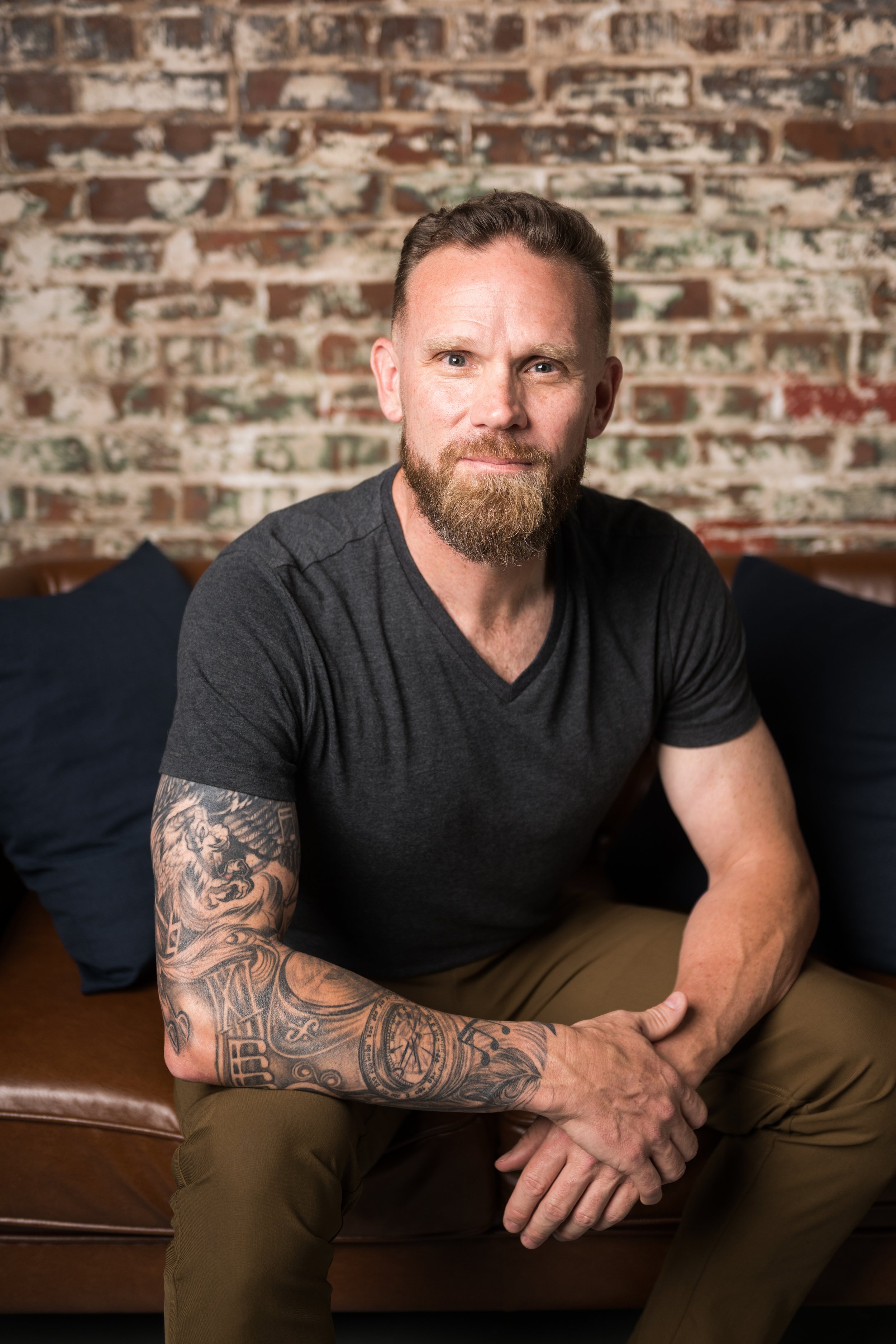 A man with a beard, tattoos on his left arm, wearing a dark T-shirt and khaki pants, sitting on a leather couch with a brick wall background.