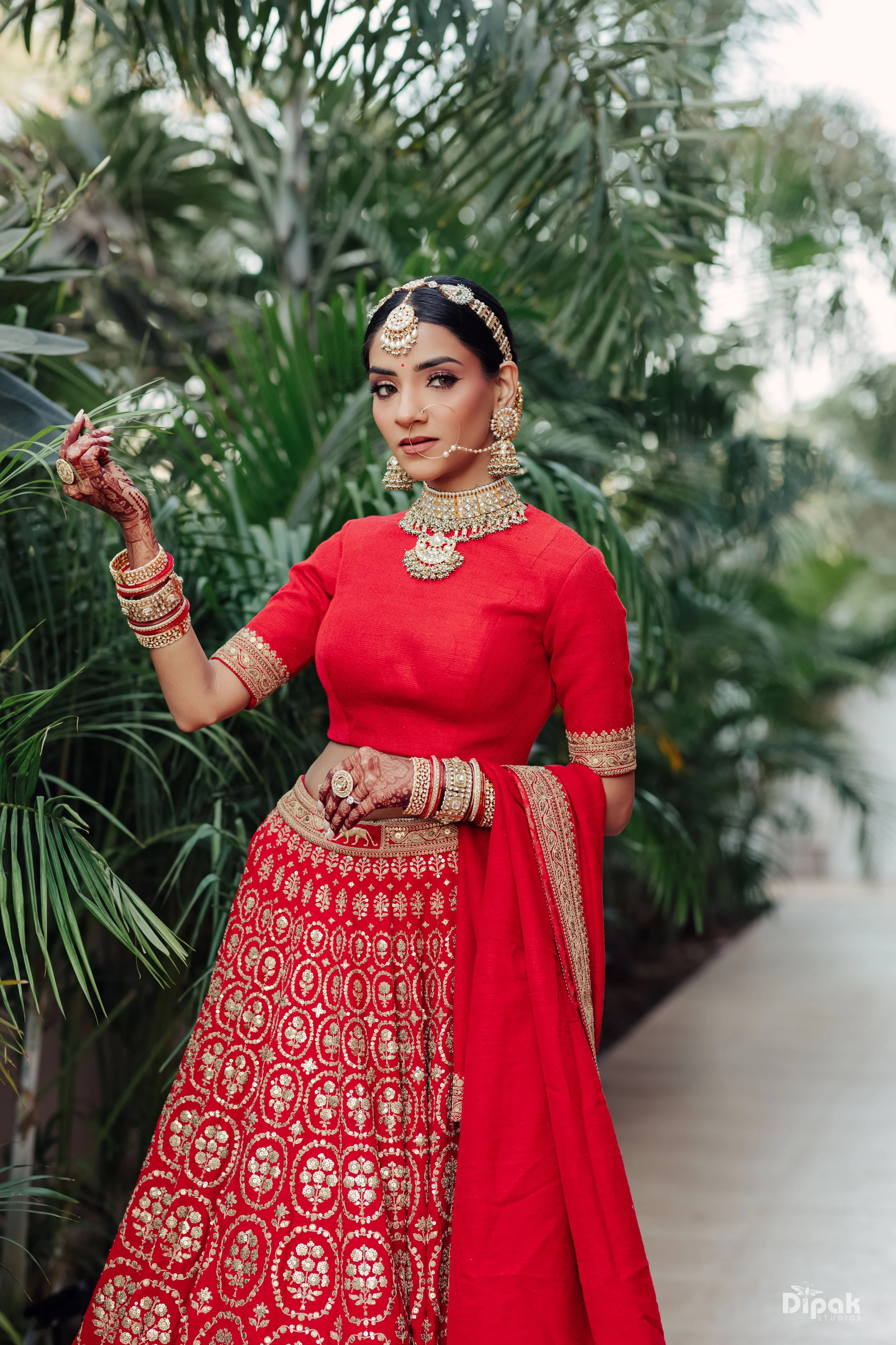 A woman in traditional Indian bridal attire, wearing red and gold embroidered clothing with heavy gold jewelry, including earrings, necklace, and bangles, standing outdoors among lush green plants.