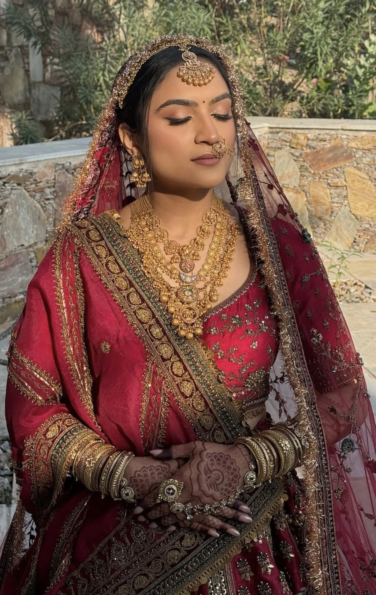 A woman dressed in traditional Indian bridal attire with elaborate gold jewelry, a red saree with gold embroidery, henna on her hands, and a maroon veil, standing outdoors with greenery and a stone wall in the background.