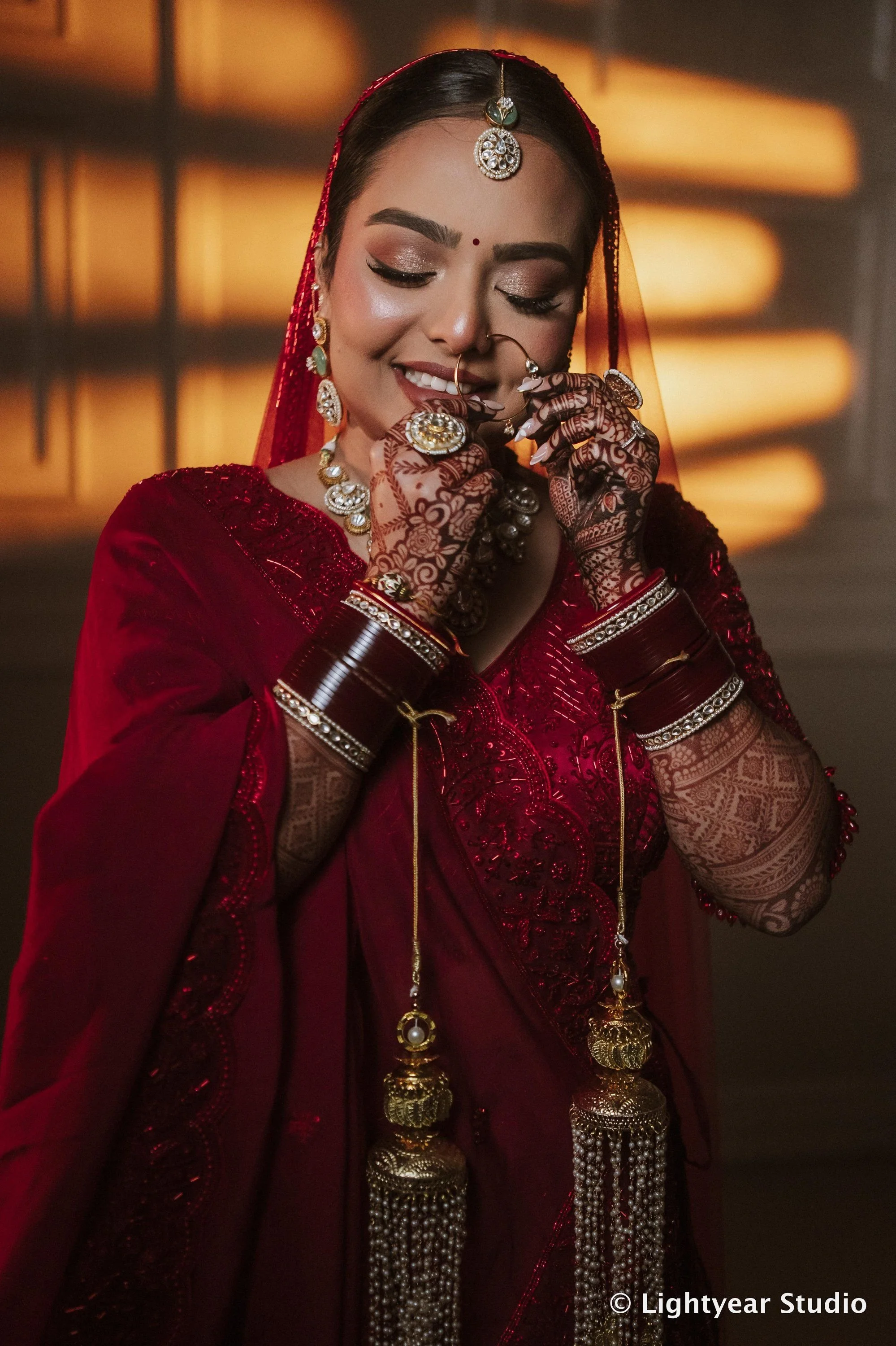A woman dressed in traditional Indian wedding attire, wearing a red dupatta, ornate jewelry, and henna decorated hands, smiling with eyes closed.