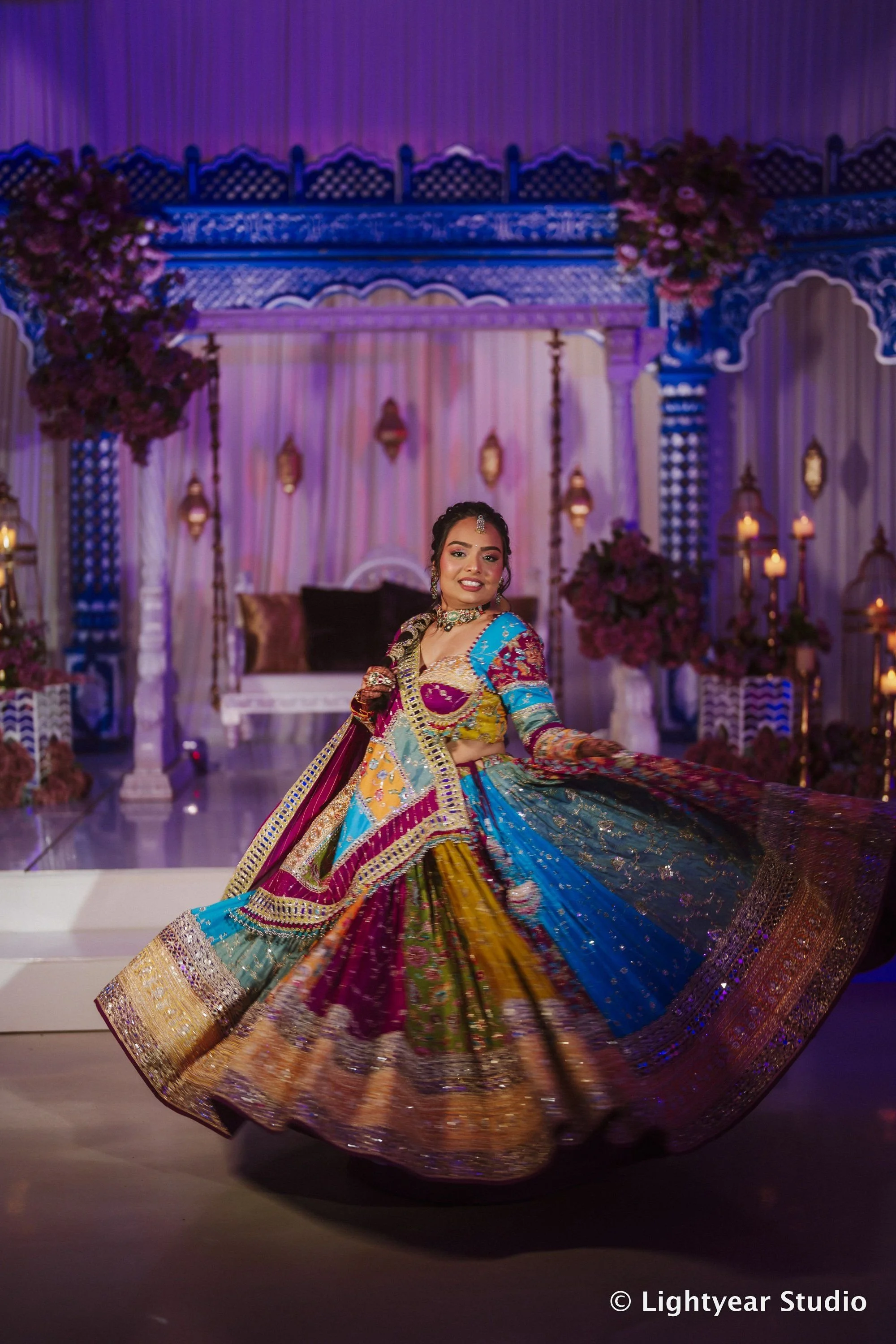 A woman dressed in colorful traditional Indian attire performing a dance on stage decorated with flowers and ornate lanterns.