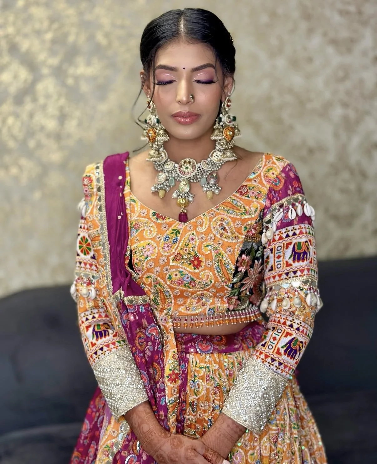 A woman dressed in colorful traditional Indian attire with intricate embroidery and mirror work, accessorized with extensive jewelry including a necklace, earrings, and a nose ring, standing with her eyes closed.