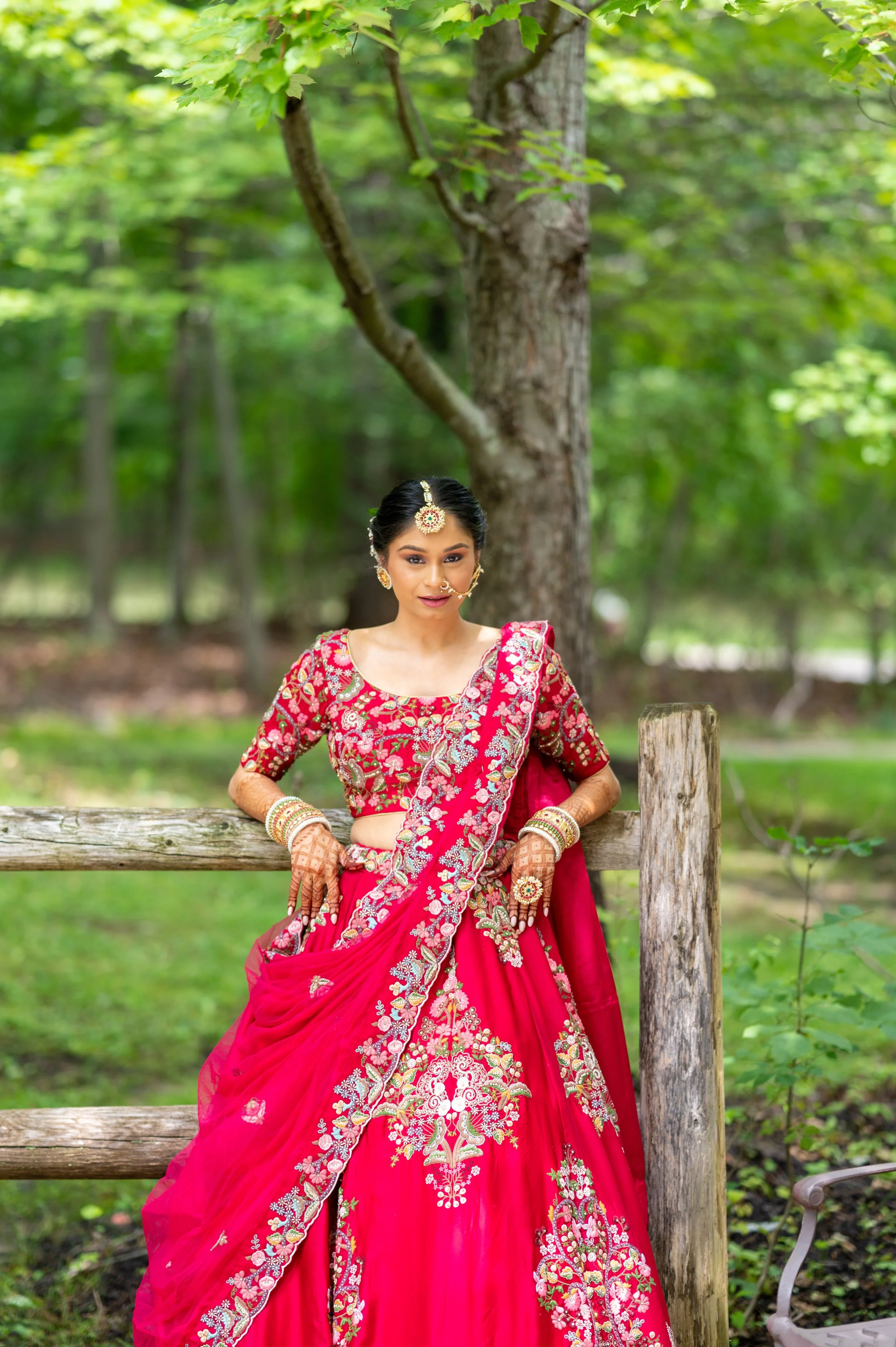 A woman dressed in a red traditional Indian bridal lehenga with intricate gold and silver embroidery, standing outdoors in a lush green forest setting.