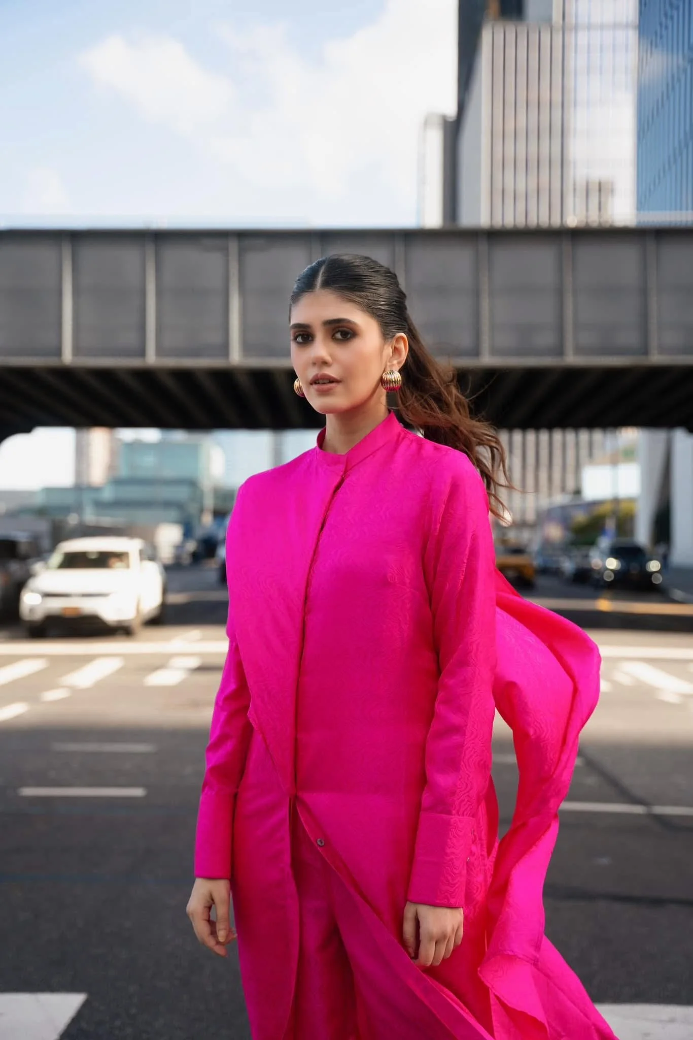 A woman wearing a bright pink traditional outfit with gold earrings, standing on a city street with a background of modern buildings and a crossing.