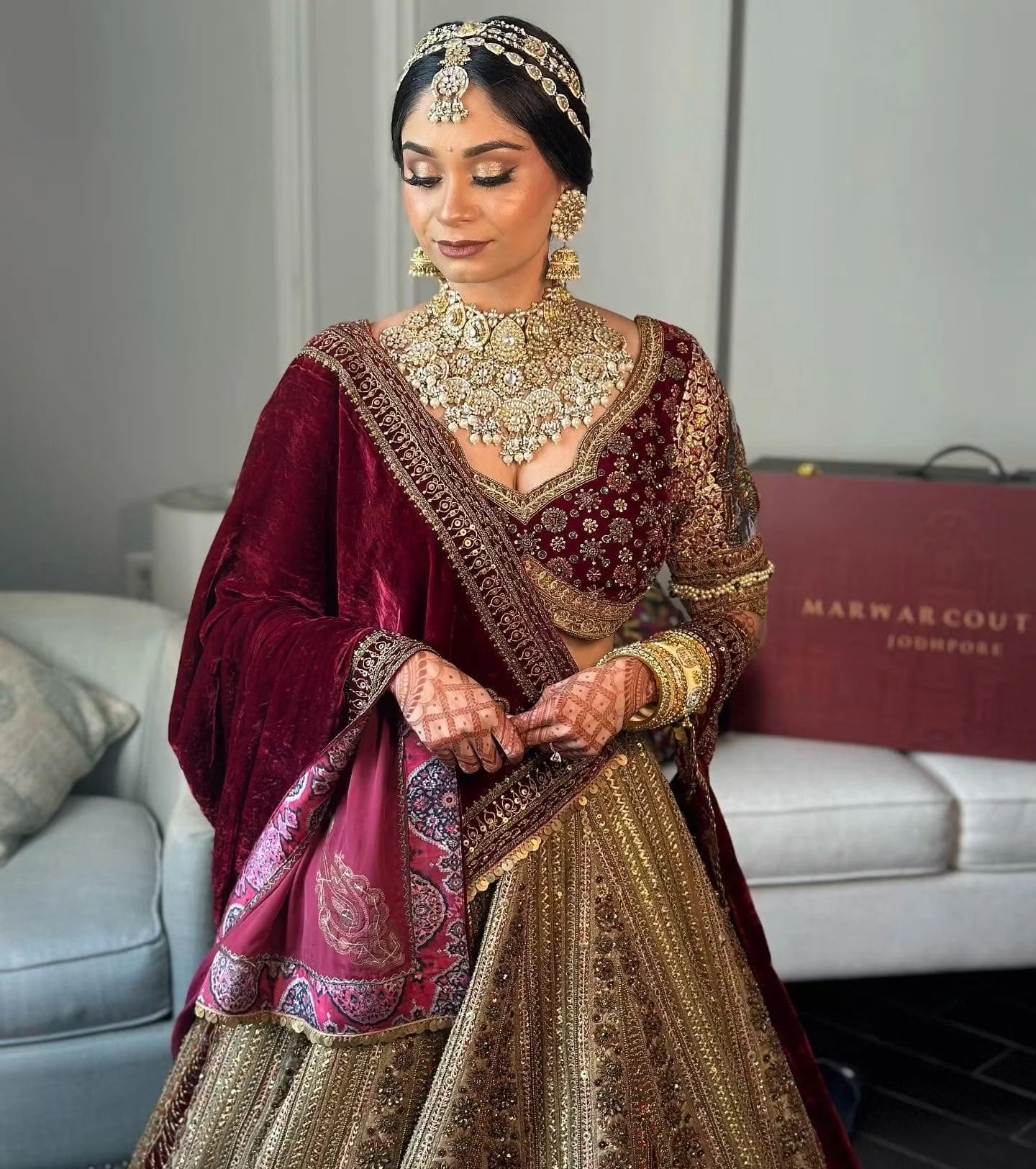 A woman wearing a traditional Indian bridal dress in deep red and gold with intricate embroidery and jewelry, including a necklace, earrings, a maang tikka, bangles, and henna on her hands.