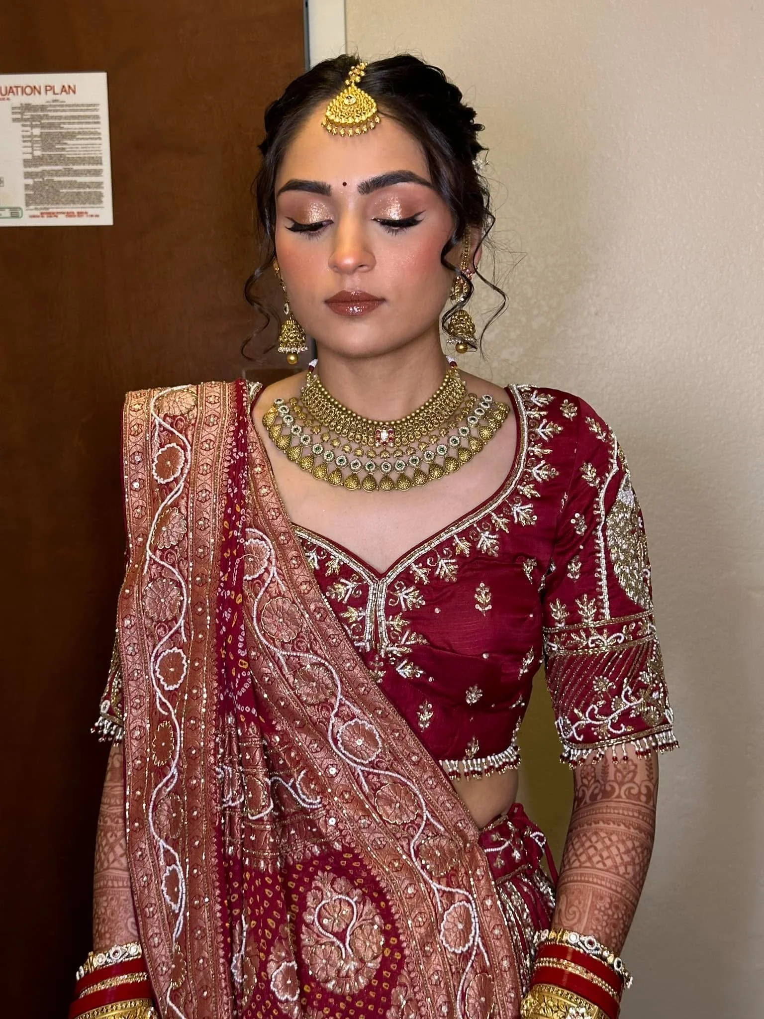 A woman dressed in traditional Indian bridal attire, wearing a red and gold embroidered saree, with elaborate gold jewelry including necklaces, earrings, a maang tikka, bangles, and henna on her hands, standing with her eyes closed.
