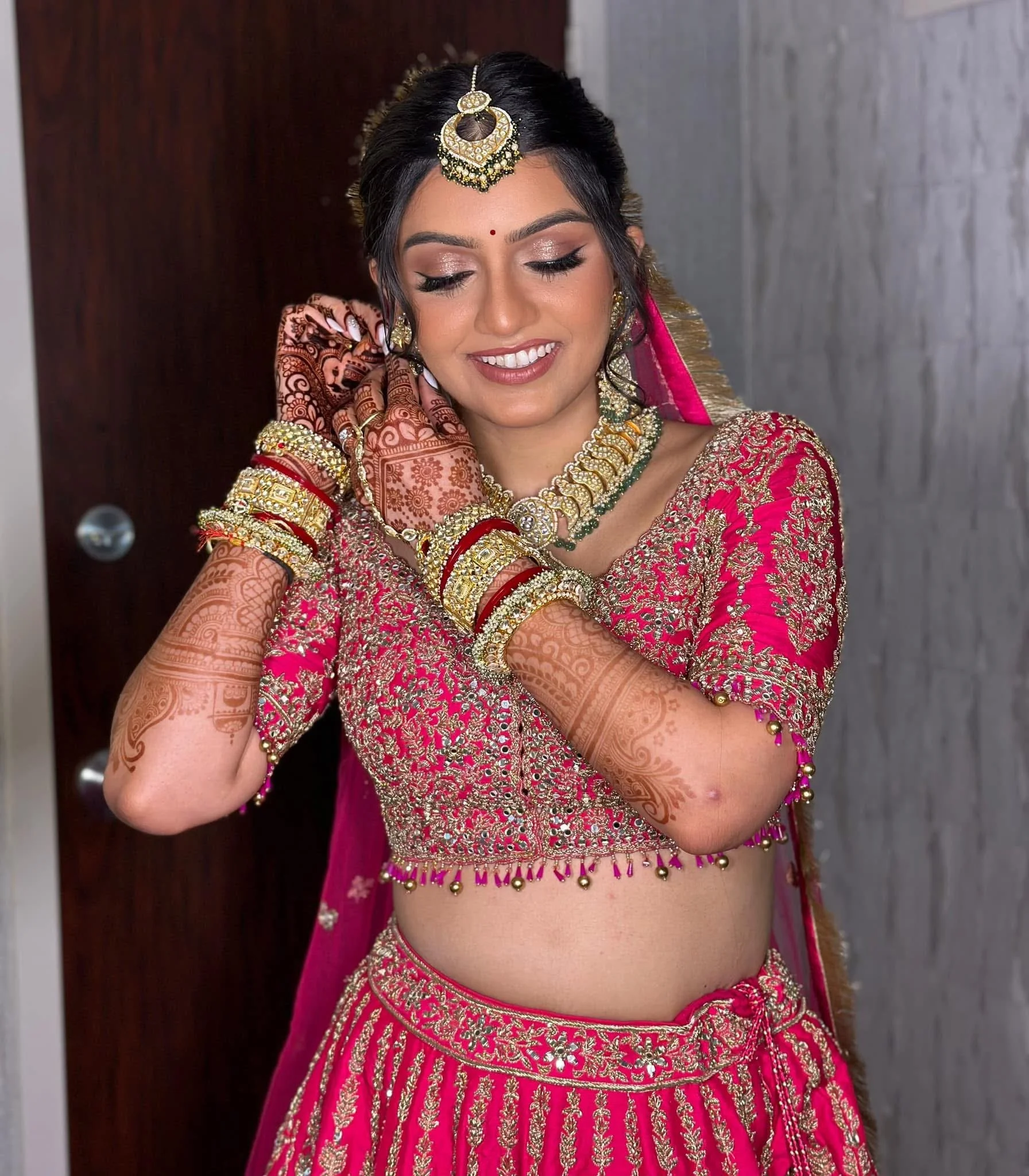 A woman dressed in traditional Indian bridal attire, wearing ornate jewelry, pink and gold embroidery, and henna designs on her arms, adjusting her earrings.