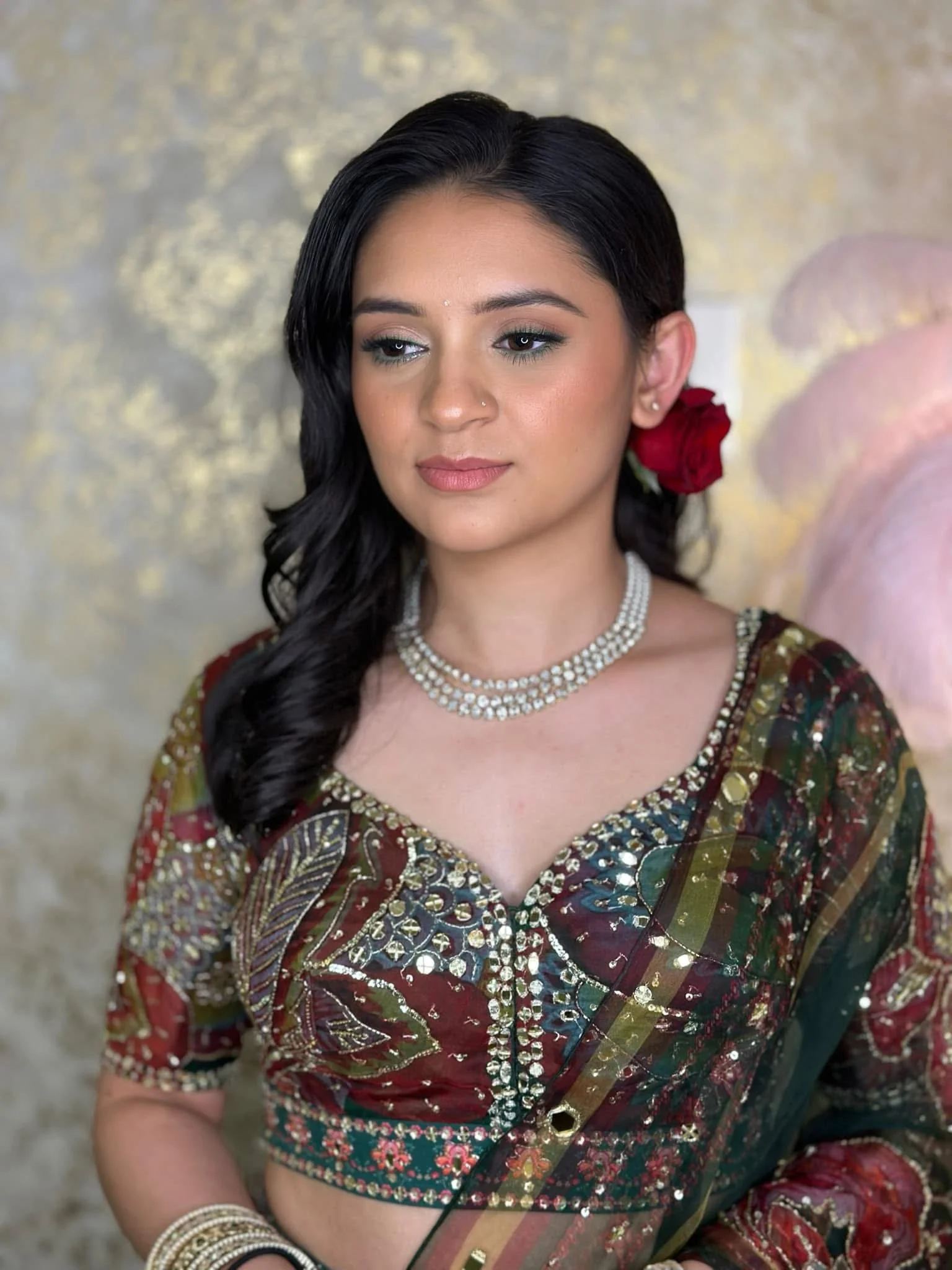 A young woman with dark hair styled in loose waves, wearing traditional Indian attire with elaborate jewelry and a red rose behind her ear, appears to be at a special occasion or celebration.