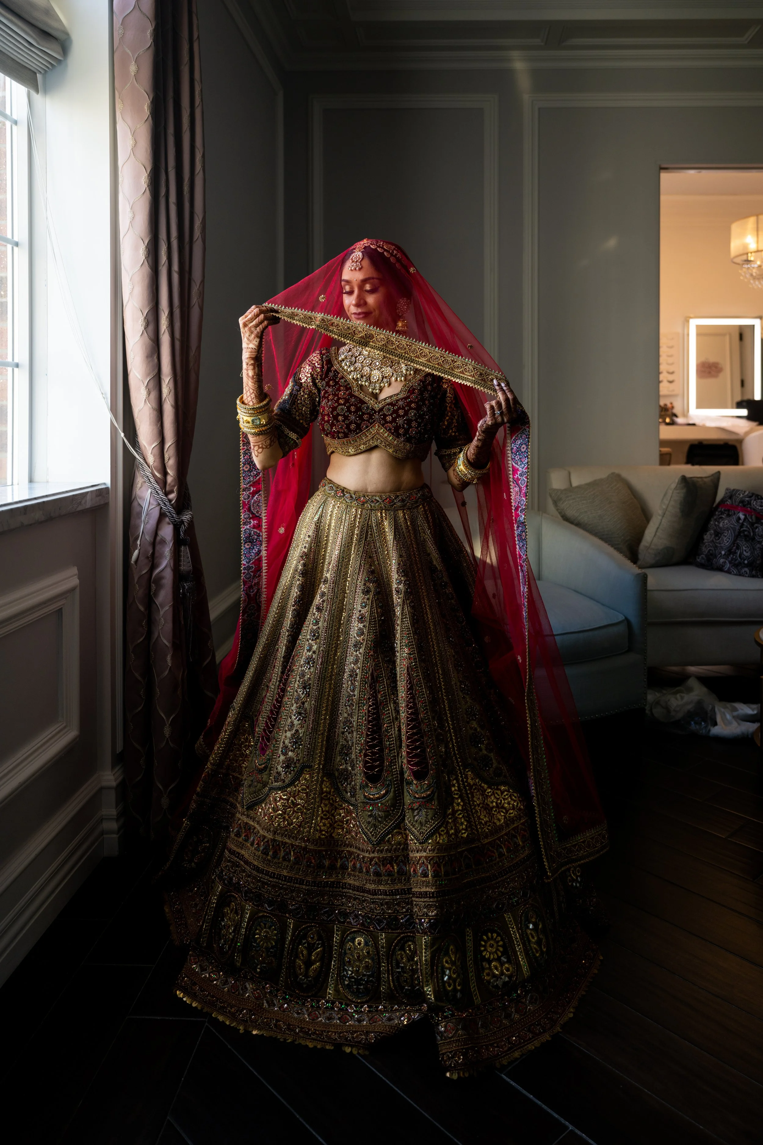 A woman dressed in an ornate traditional Indian bridal outfit, including a heavily embroidered lehenga, choli, and dupatta, standing in a room near a window with curtains, adjusting her dupatta.